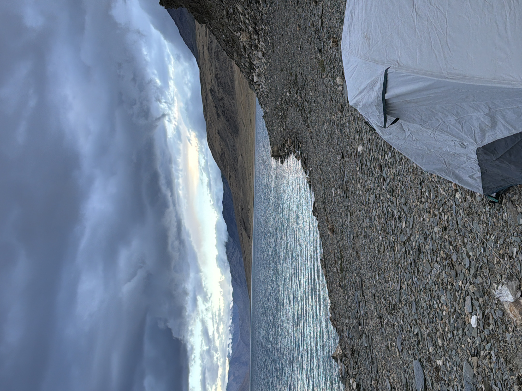 The image depicts a campsite on the rocky shore of a large, choppy lake in a mountainous region. A white tent with a grey base is pitched in the foreground on the right side of the frame. The lake occupies the midground, its surface rippled by wind, reflecting the overcast sky. In the background, rolling, barren hills rise up towards a mountain range shrouded in clouds. The sky itself is filled with dramatic, dark, and grey clouds, with hints of lighter sky breaking through in the distance. The overall scene suggests a remote and rugged natural environment.