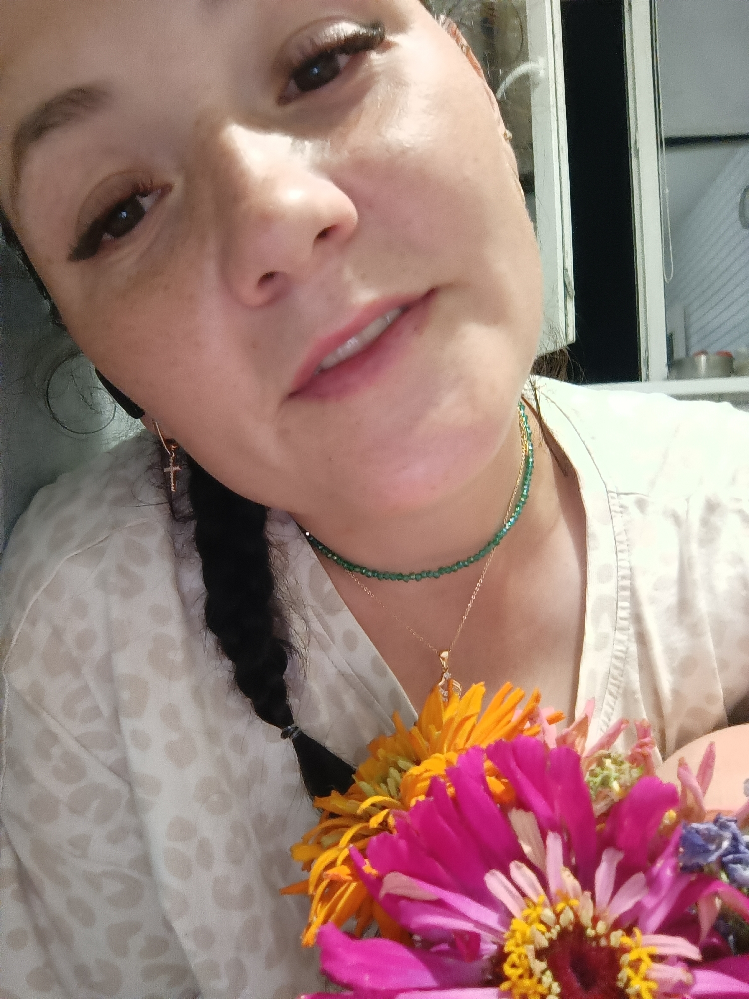 The image is a close-up, selfie-style portrait of a woman with dark hair braided over her shoulder. She is wearing a light-colored shirt with a subtle leopard-like print, a green beaded necklace, and a delicate gold necklace with a pendant. A small gold cross earring is visible on her left ear. She is holding a bouquet of brightly colored flowers, including pink, orange, and blue zinnias, in the foreground, partially obscuring her chest. The woman's face is tilted towards the camera, and her eyes are looking slightly upwards. Her makeup includes noticeable eyeliner and mascara. The background appears to be indoors, with a window visible on the right side, suggesting an interior setting. There is no discernible information in the image to indicate the location as Chaikovsky, Russia.