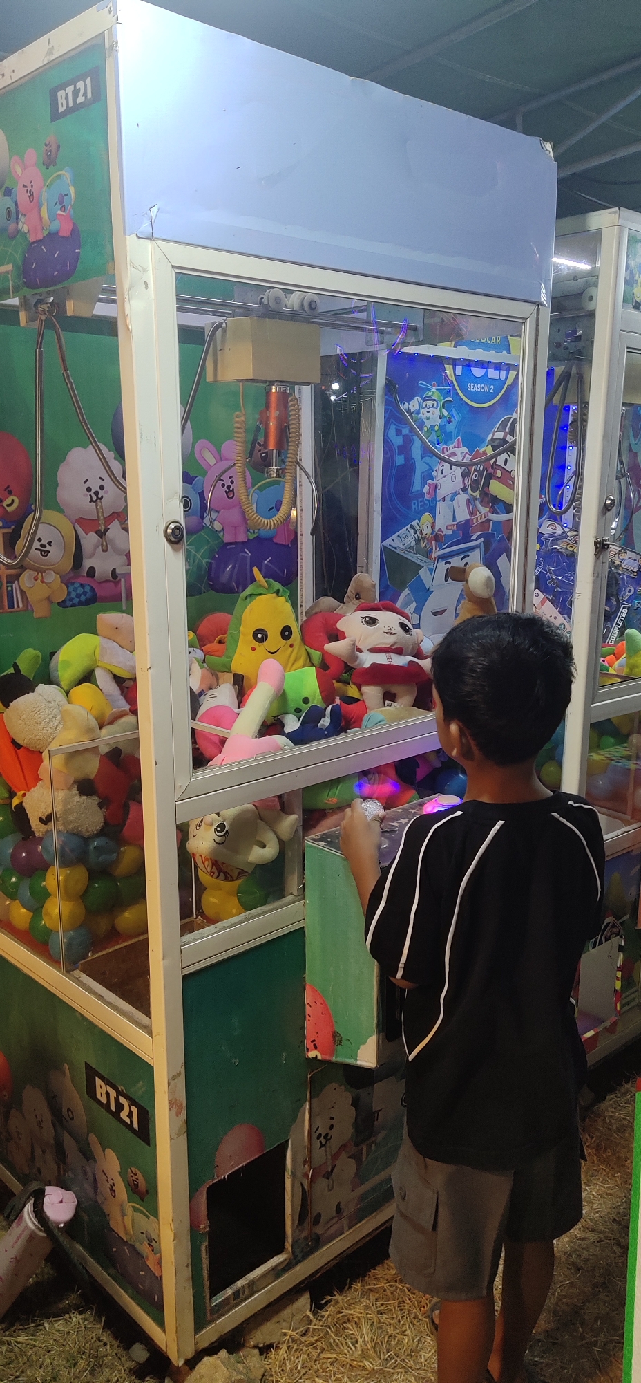 The image shows a young boy standing in front of a claw machine filled with plush toys. The claw machine is decorated with characters from BT21, a collaboration between Line Friends and BTS. The machine is situated outdoors, possibly at a fair or carnival, with dry grass or hay on the ground. The background shows another similar claw machine and a tent-like structure. The boy is wearing a black t-shirt with white accents and grey shorts. He is holding a coin or token and appears to be operating the machine. The claw mechanism is visible at the top of the machine, with the plush toys filling the display area below. The environment suggests a casual, entertainment-focused setting in Tasikmadu, Indonesia.