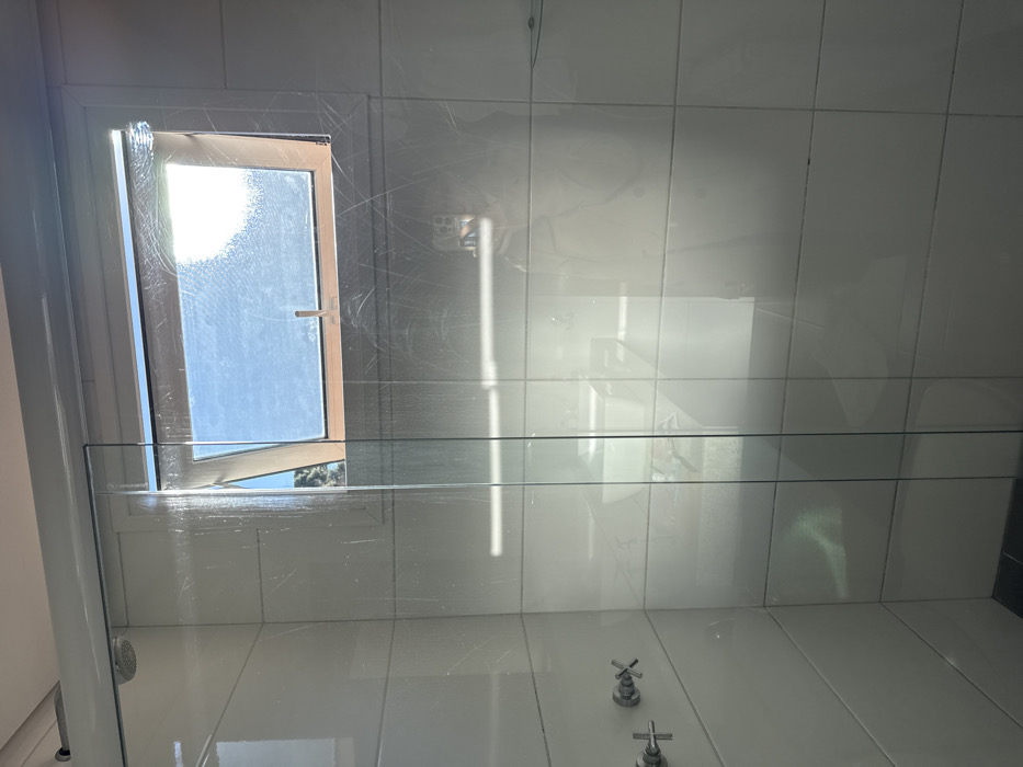This forensic description details a bathroom shower area located in Canela, Brazil. The scene is dominated by a transparent glass shower enclosure, which features numerous visible water streaks, smudges, and faint scratches across its surface. To the left, within the shower, the walls are covered in rectangular white tiles, upon which a chrome-finished shower head is mounted in the upper left corner, and two chrome cross-shaped faucet handles are visible on the lower left.
Above the shower enclosure, a top-hinged window with a light-colored frame is partially open, revealing a clear blue sky and distant green foliage, indicating an outdoor view. Bright sunlight streams through this window, creating strong reflections on the glass. Through the glass, on the lower right side, a reflection of a white bathroom sink or vanity with a faucet is discernible, positioned behind the camera's viewpoint. A clear reflection of a person holding a smartphone, the likely photographer, is also evident on the right side of the glass enclosure. The floor within the shower appears to be a dark color. No other individuals or direct interactions are visible in the scene.