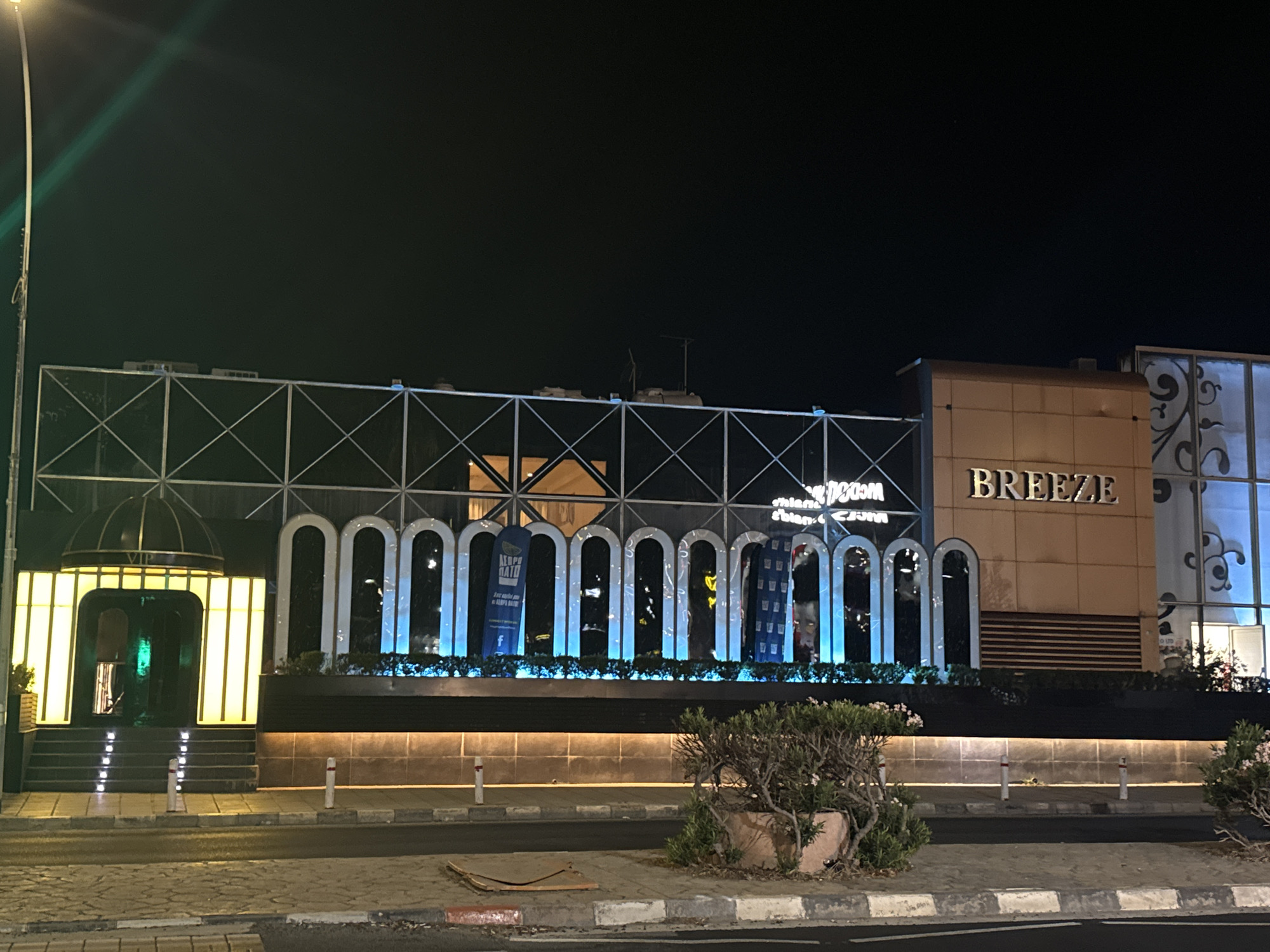 This is a night-time, eye-level, medium shot of the entrance to a building identified by the sign "BREEZE". The building facade is lit with a combination of warm yellow lights illuminating an arched entrance with stairs, and cool blue lights highlighting a series of arched windows or doorways. A street lamp with a green light is visible on the left side of the frame. The building appears to be a commercial establishment, possibly a club or restaurant, given the signage and illuminated design elements. The surrounding environment includes a paved sidewalk, a road, and a street with white bollards. A small, dry bush in a planter is in the foreground on the right. The sky is dark, indicating it is night. No people are visible in the image. The location is identified as Germasogeia Municipality, Cyprus.