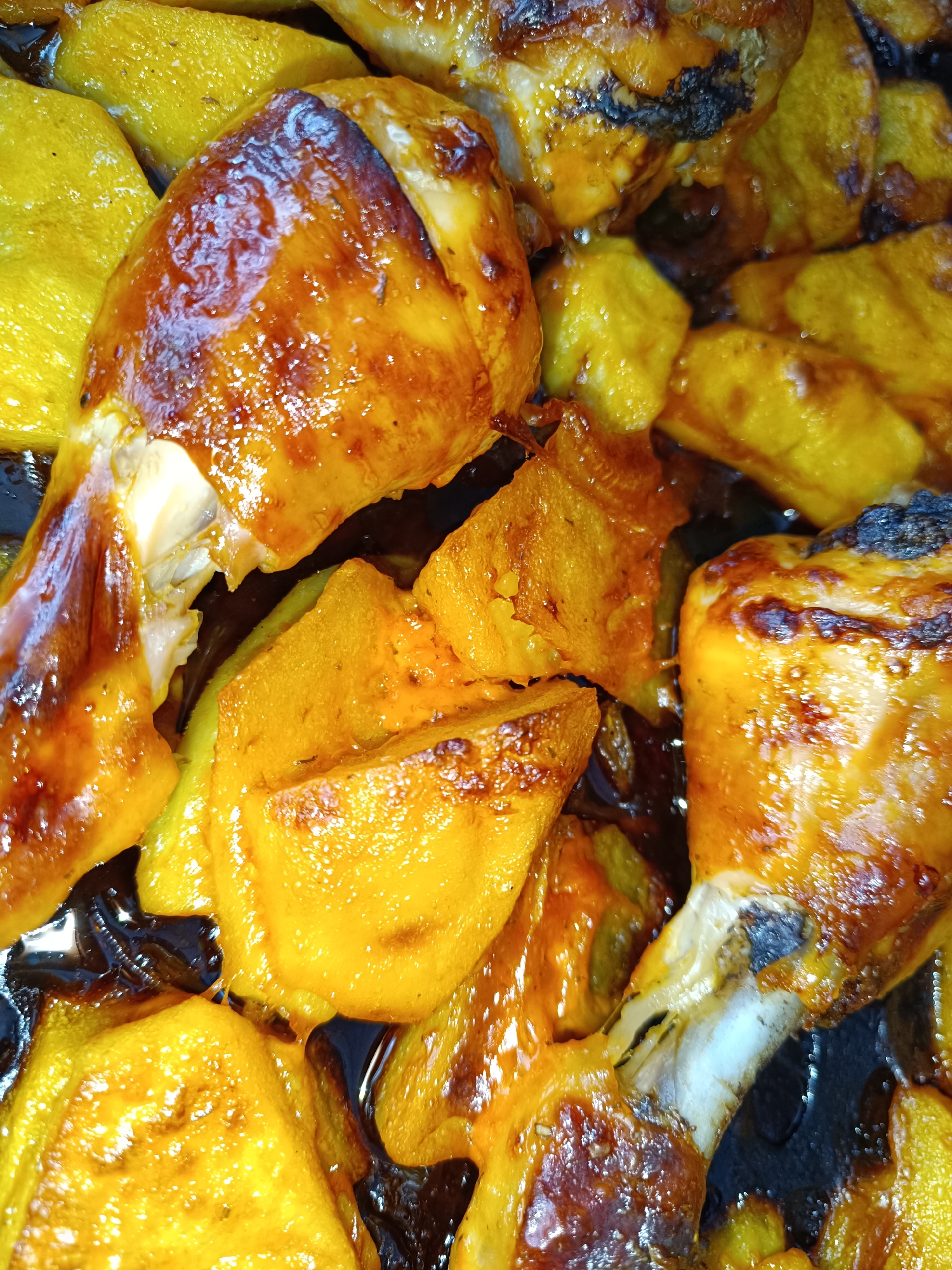 The image depicts a close-up shot of glazed chicken drumsticks mixed with what appears to be cooked yellow squash or pumpkin. The food is in a dark pan or dish, with a shiny, dark sauce pooling at the bottom. The lighting is bright and highlights the glaze on the chicken and squash, emphasizing the textures. The image does not provide information to determine the location in the city of Tobolsk, Russia. There is no visible information about people, actions, or interactions.