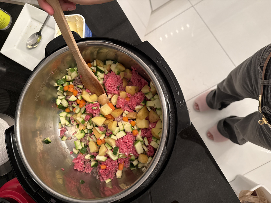 A top-down shot shows an Instant Pot filled with ingredients: ground meat, diced zucchini, potatoes, and carrots. A wooden spoon stirs the mixture. Nearby, a white dish holds a spoon with a dab of something creamy, possibly butter, and a tub of butter. A person wearing gray pants and pink slippers stands beside the pot. The scene suggests food preparation, possibly making a stew or soup in a modern kitchen.