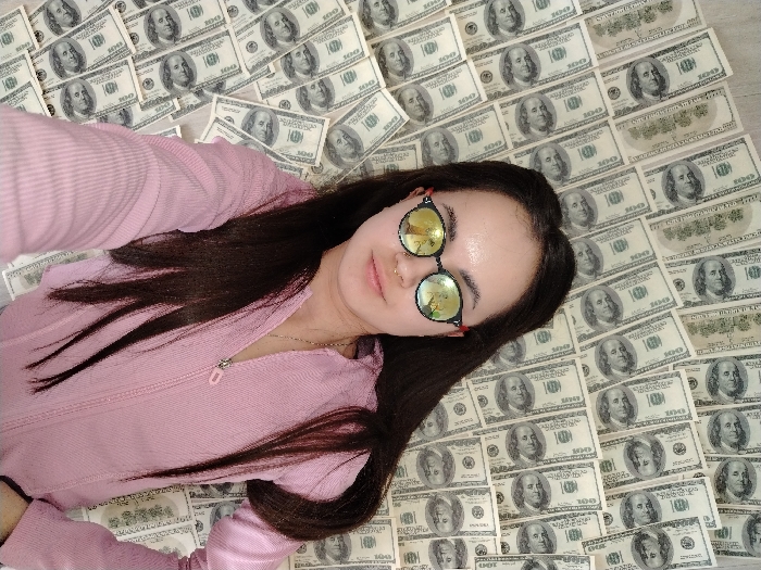 A female individual is depicted lying prone on a surface completely covered with US one-hundred dollar bills. The subject has long, dark hair and is wearing a light pink, ribbed, zippered jacket. She is also wearing round, reflective sunglasses with red frames and a small piercing on her left nostril. Her head is positioned towards the upper right, and she appears to be looking directly at the camera. Her left arm is visible, extending towards the lower left. The currency is spread out in an overlapping pattern, forming the entire background of the scene. The specific location in the city of Noviy, Russia, cannot be determined from the visual information provided in the image.