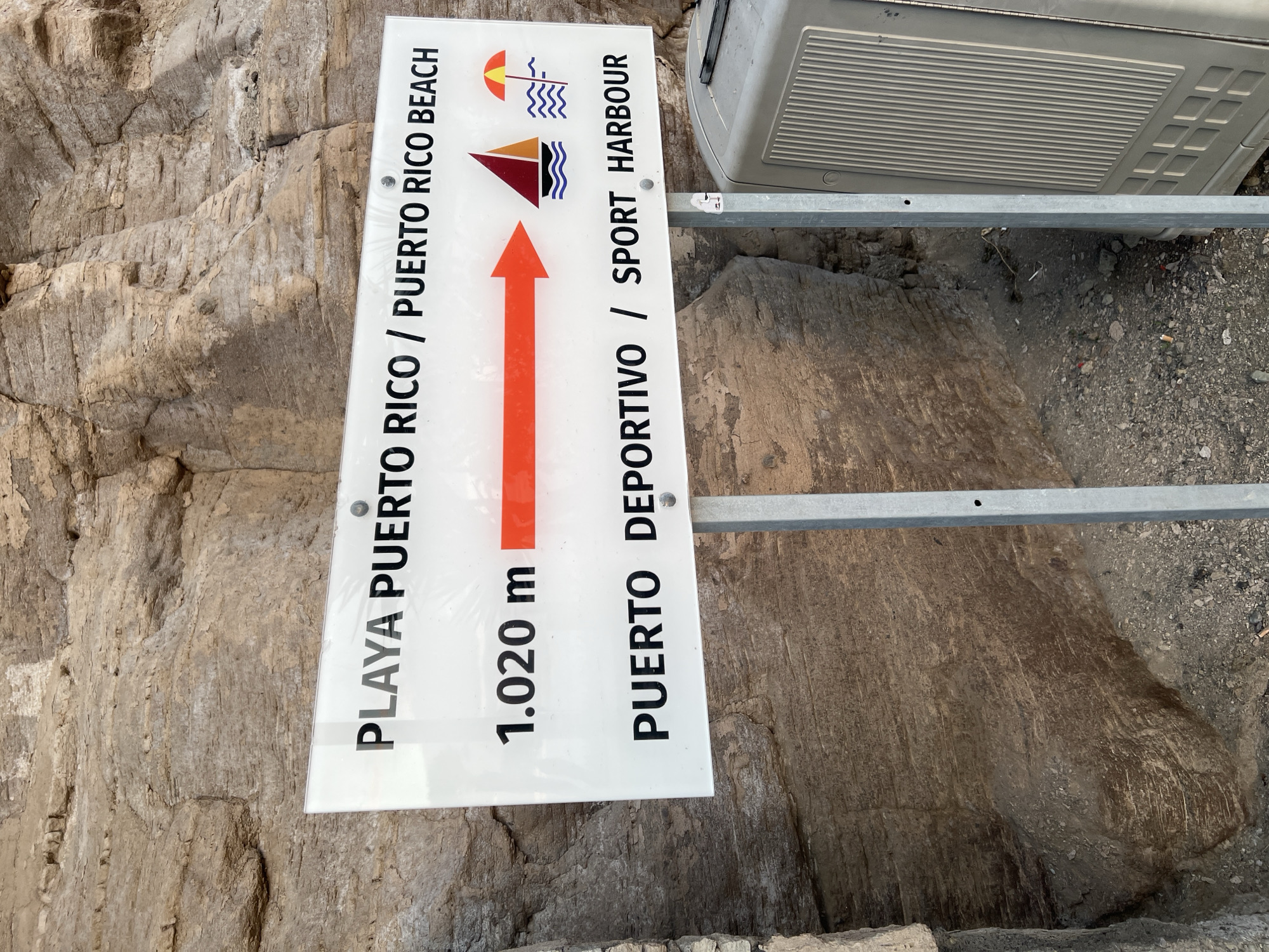 The image shows a directional sign in Mogán, Spain, indicating that the Playa Puerto Rico beach and the Puerto Deportivo sport harbor are 1.020 meters in the direction indicated by the red arrow. The sign is attached to a metal frame. The background shows a rocky terrain.
