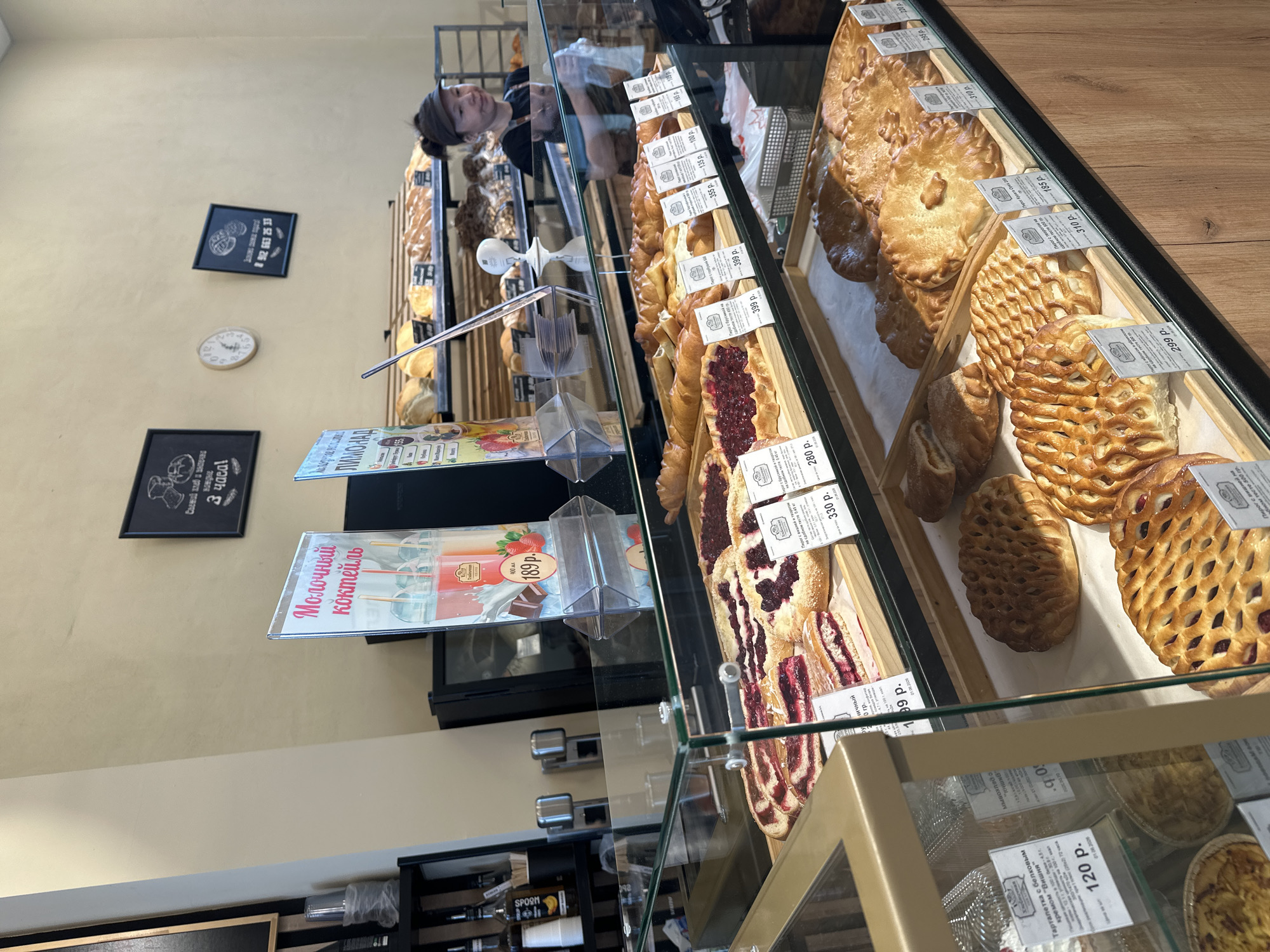 An indoor, eye-level photograph depicts a bustling bakery in Yekaterinburg, Russia, identified by a local phone number (8 912 663 25 33) on a wall sign and prevalent Russian text. The central focus is a multi-tiered glass display case filled with a diverse array of freshly baked goods.
On the top tier of the display, various large pies and tarts with fruit fillings, predominantly red and purple berries, are arranged on wooden trays. Items include a cherry and curd pie (330 rubles) and a lingonberry pie (280 rubles), among others with prices ranging from 280 to 399 rubles. The lower tiers showcase other pastries, some with braided or lattice tops, and large savory pies, such as a chicken-mushroom pie (185 rubles), with prices generally between 185 and 310 rubles. White price tags in Russian rubles are placed next to each item.
Behind the display cases, a woman of East Asian or Southeast Asian descent, wearing a dark cap and apron, stands behind a dark countertop. Her gaze is directed towards the right side of the frame. Behind her, shelves are stocked with various loaves of bread and other baked items. To the far left, a shelf unit holds bottles of syrups, plastic cups, and cup dispensers.
Two vertical promotional standees are positioned on the counter: one advertising "Молочный коктейль" (Milkshake) for 189 rubles, featuring vibrant drink images, and another for "ЛИМОНАД" (Lemonade) for 155 rubles. The background wall, painted a light beige, features three framed elements: a black sign stating "Свежий хлеб в пекарне каждые 3 часа!" (Fresh bread in the bakery every 3 hours!), a round analog clock displaying approximately 12:15, and the aforementioned black sign with the local phone number. The bakery is brightly lit, presenting a clean and organized commercial environment.