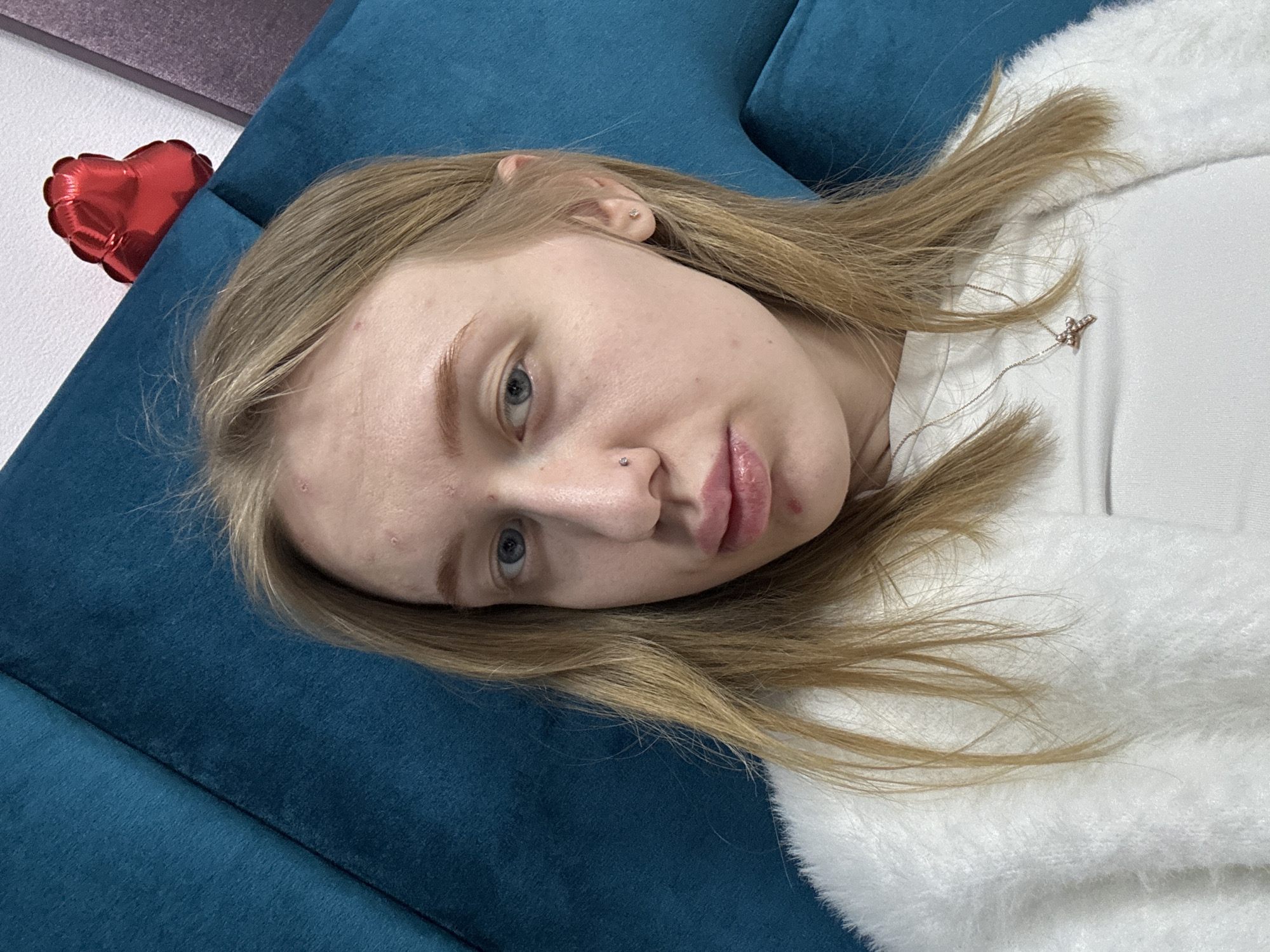 A fair-skinned young woman with light blonde hair lies on a blue cushioned surface, likely a sofa. She wears a white fuzzy garment. The background includes a white wall with a red heart-shaped balloon. No information indicates the scene is in Rostov-on-Don, Russia.