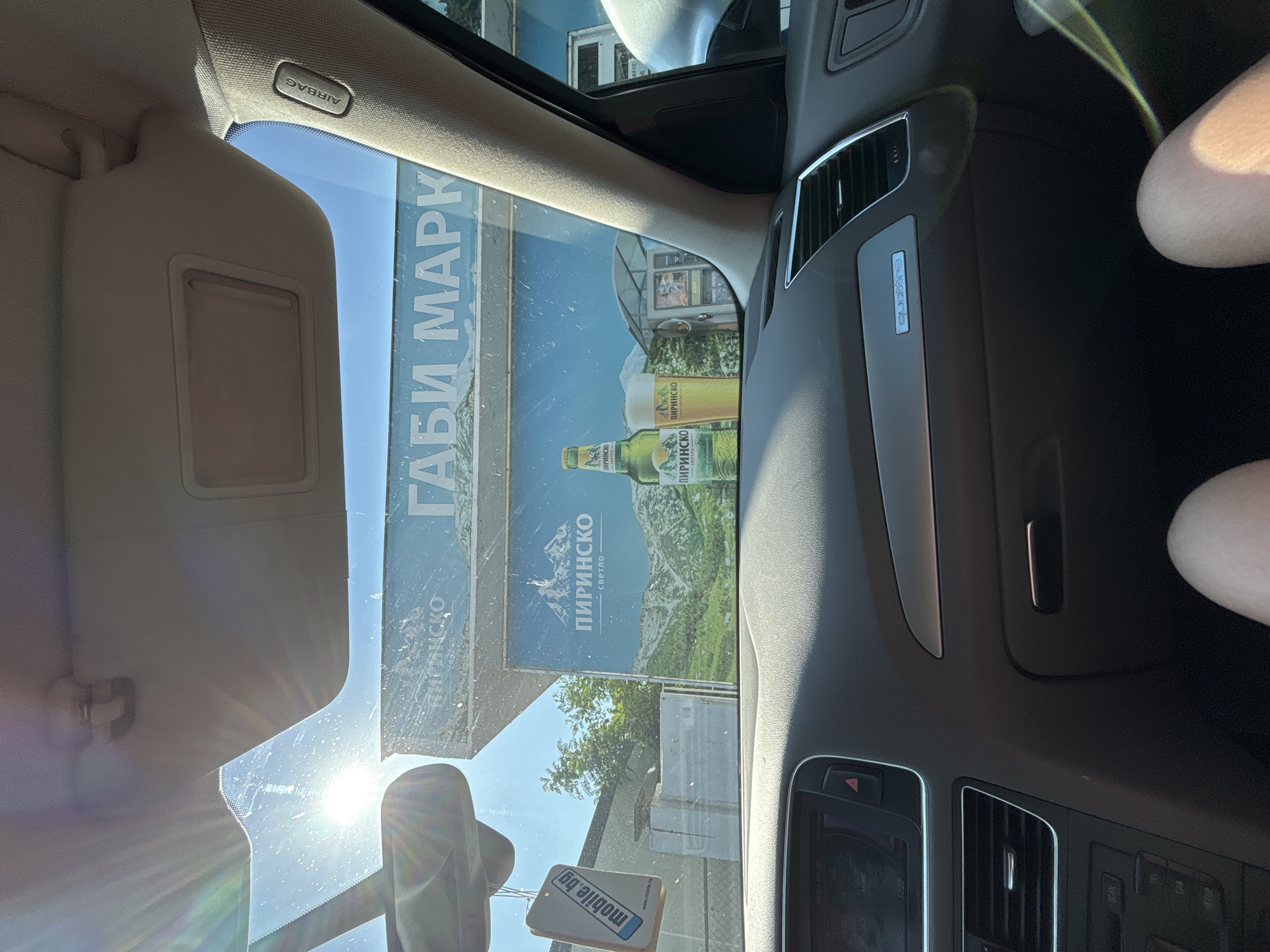 An eye-level shot, likely taken by a passenger, looks out from the interior of a car through a sun-drenched windshield. The image is oriented vertically (rotated 90 degrees clockwise from typical viewing).
Inside the car, the beige sun visor is visible on the upper left, bearing an "AIRBAG" label. Part of the car's dashboard, air vents, and central console are seen on the right, featuring a "Quattro" badge and a digital display showing "1:20". Below, several buttons are visible including "SCAN", "TEL", "CAR", and "SETUP", along with a hazard light button. A "mobile.bg" air freshener hangs from the rearview mirror in the lower center. A bare human leg is partially visible on the far right. The windshield has some smudges and significant lens flare from the bright sun.
Outside the car, the primary subject is a large billboard advertising "ПИРИНСКО" (Pirinско) beer, a Bulgarian brand, with text "светло" (svetlo - light). The billboard also displays "ГАБИ МАРК" (Gabi Mark) and features an image of mountains and beer bottles/glasses. The background reveals a clear blue sky, some green trees, and parts of buildings. The bright, clear weather suggests it's daytime in Rakovski, Bulgaria, as indicated by the context and Bulgarian text.