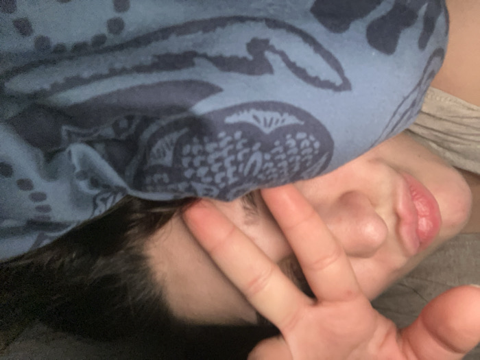 A person's face is partially visible under a blue patterned cloth. The face is tilted, with lips slightly pursed. A hand displaying a peace sign is positioned in front of the face, partially obscuring the nose and one eye. Dark hair is visible near the forehead. A gray garment is also partially visible. The overall impression is of someone playfully concealing their face.