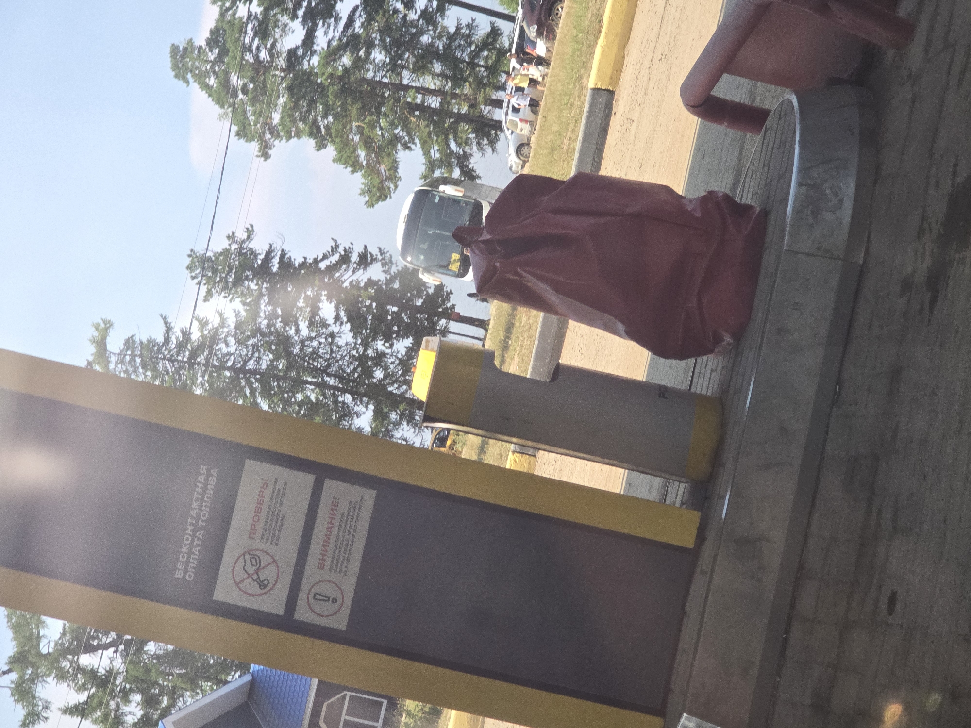 The image depicts a street scene in Turka, Russia, on a sunny day. In the foreground, a red plastic bag rests on a paved surface, possibly at a gas station or a public rest area, next to a yellow and gray dispensing pillar. The pillar has signs with Russian text, including one that reads "Бесконтактная оплата топлива" (Contactless fuel payment). Further back, a white bus is parked, and several cars are visible. The background features a body of water, likely a lake or river, and lush green trees. People are seen standing near the bus and cars, suggesting a gathering or a stop during a journey. The overall atmosphere is casual and relaxed, with clear skies and bright sunlight.