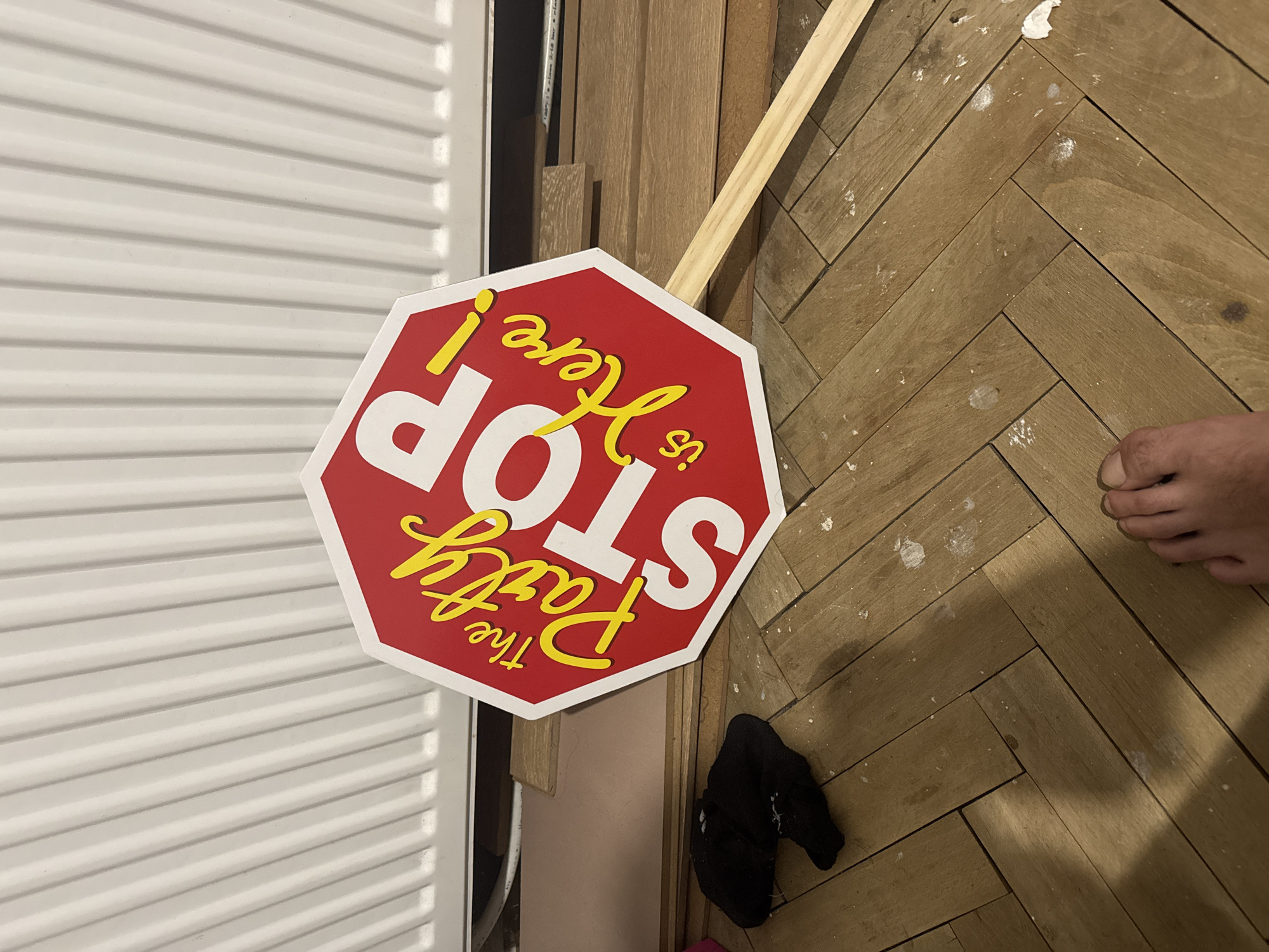 A stop sign that reads "The Party is Here!" is lying on its side on a wooden floor. The sign is red with white and yellow lettering and has a wooden handle. A black sock is on the floor to the left of the sign. A person's bare foot is visible in the lower right corner of the image. The floor appears to be made of parquet flooring. In the background, a white radiator is visible. The scene appears to be indoors, possibly in a residential setting. There are no visible landmarks or indications of the city of Sofia, Bulgaria.
