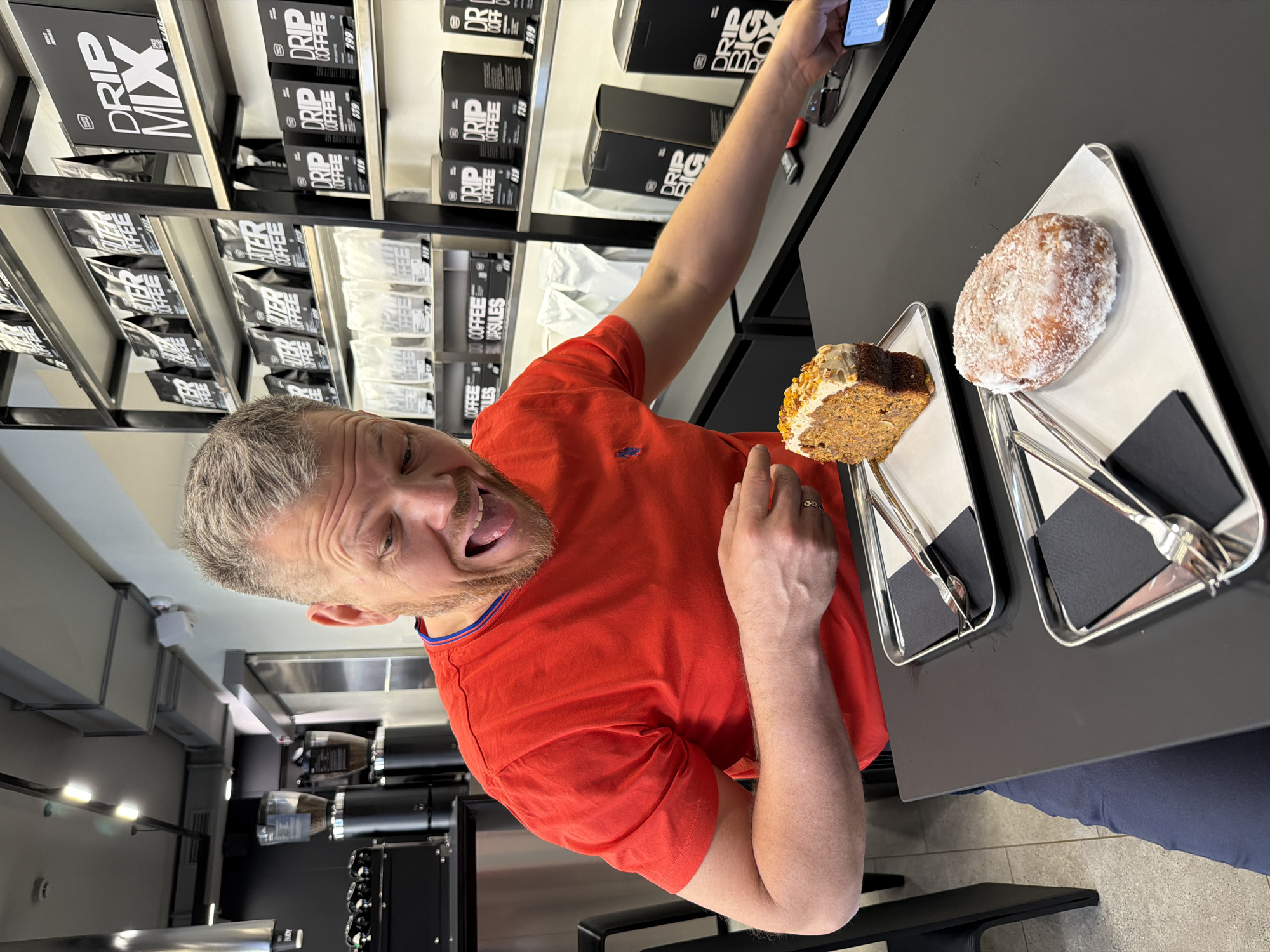 The image captures a person in what appears to be a coffee shop, judging by the shelves filled with "Drip Coffee" and "Filter Coffee" products. The individual is wearing an orange t-shirt and is holding a smartphone as if taking a selfie, making a funny face with their tongue out. On the table in front of the person, there's a piece of cake and a powdered donut on individual trays, each with utensils. The shop has a modern interior, indicated by the lighting, the black tables, and what appears to be a commercial coffee machine in the background. The location is not specified in the image, so it is not possible to say it is in Izhevsk, Russia.