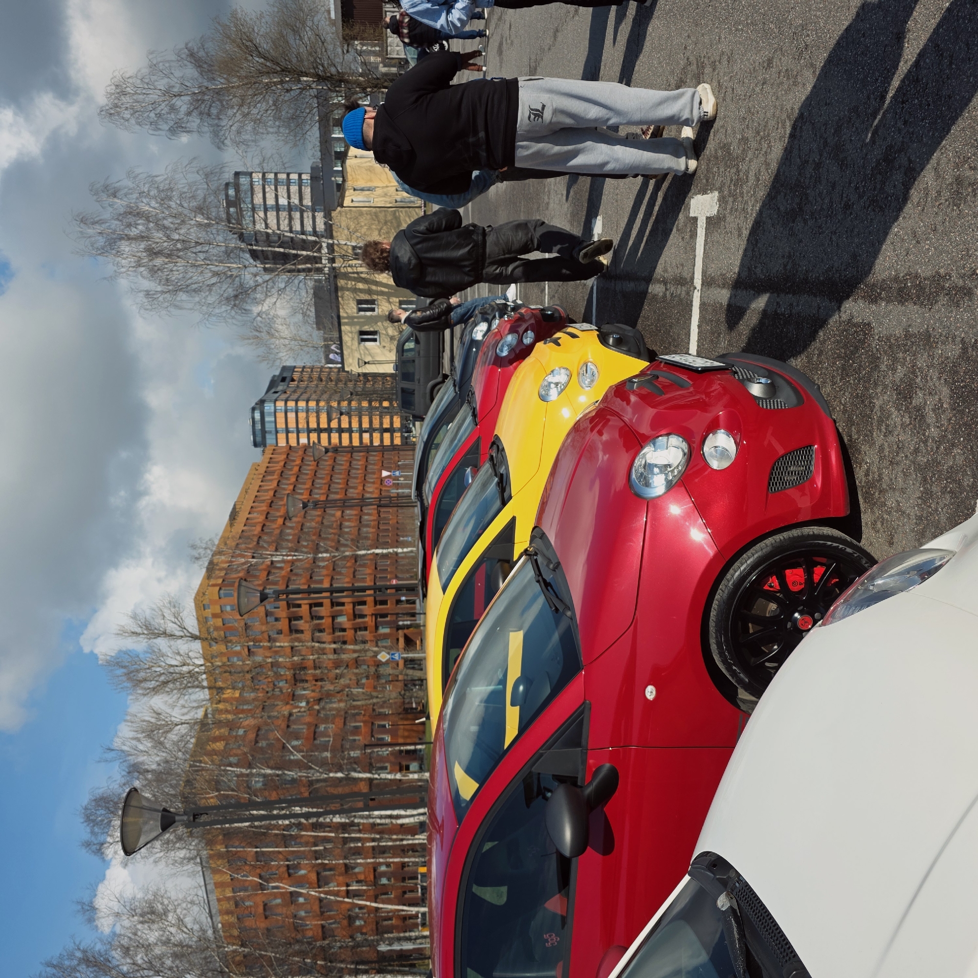 The image captures a parking lot scene in Saint Petersburg, Russia, filled with a row of colorful Fiat 500 Abarth cars, dominated by red and yellow hues. Several individuals are visible: one wearing a blue hat, black jacket, and gray sweatpants, another in a black leather jacket, and others further back, their details less clear. These people are standing in the parking lot next to the cars. The vehicles are lined up neatly within parking spaces marked by white lines. The backdrop features modern buildings with brick and glass facades under a partly cloudy sky. Bare trees are scattered throughout, suggesting a cooler season. A street lamp is visible in the background. The overall setting is urban, with a mix of architecture and natural elements.
