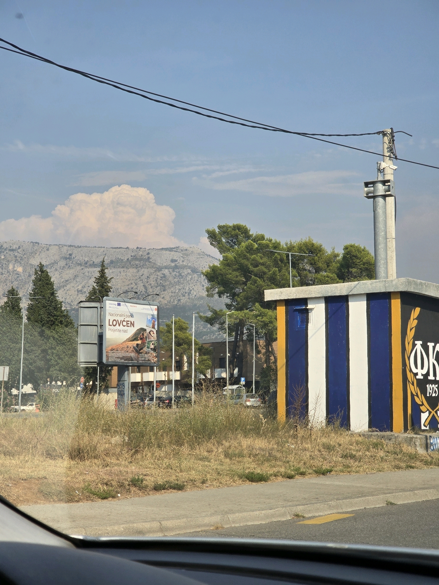 The image depicts a scene in Podgorica, Montenegro, on a sunny day. In the foreground, dry, yellowed grass lines a gray asphalt road with a yellow painted line. A sidewalk separates the road from a patch of dry vegetation. Behind the vegetation, a small, rectangular building is visible, painted with vertical stripes of white and royal blue, bordered by a wide yellow stripe on the right side. On this yellow stripe, the Greek letters "ΦΚ" are painted in black, along with the year "1925" below. A laurel wreath emblem is also depicted.
To the left of the building, a large billboard stands on a metal pole. The billboard advertises "Nacionalni park LOVCEN - Posjetite nas!" (National Park Lovcen - Visit us!) and features images of people enjoying the park. In the background, a line of trees, including evergreens and deciduous trees, partially obscures a mountain range under a bright blue sky with scattered clouds. Power lines stretch across the sky, supported by a utility pole. The overall impression is an outdoor scene in a semi-urban or rural area.