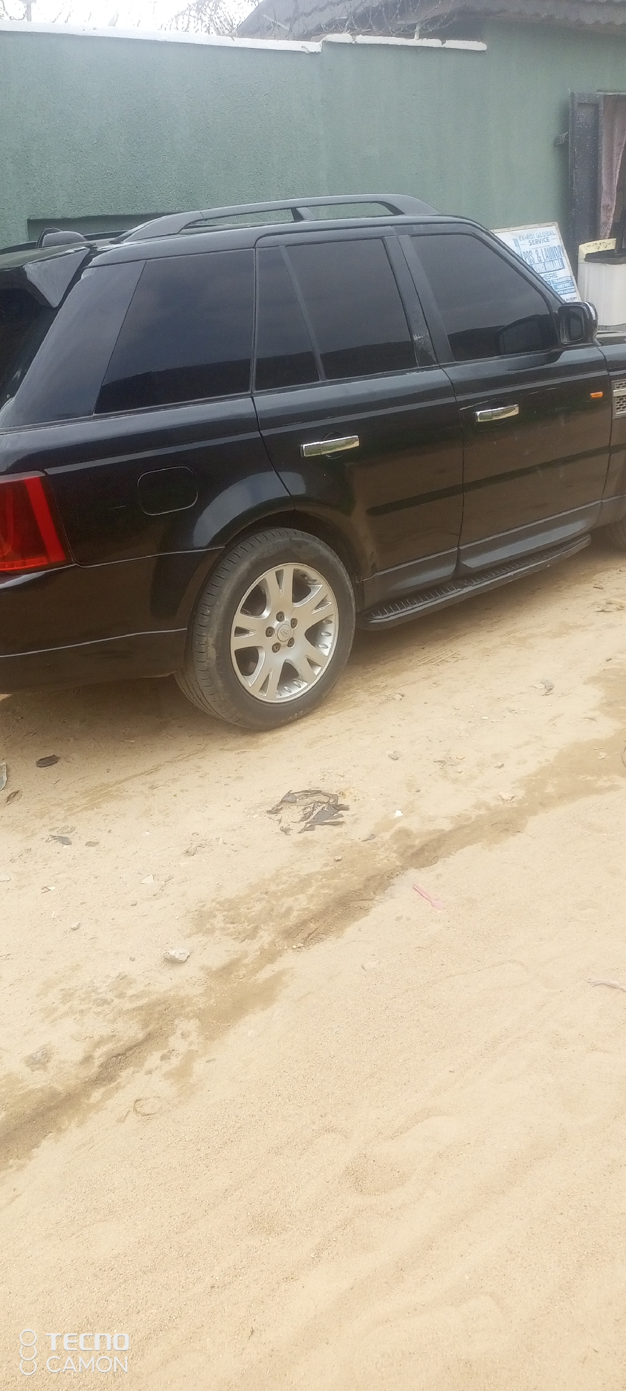 A black Range Rover Sport SUV is parked on a dirt lot in Jalingo, Nigeria. The vehicle is facing right, and its windows are tinted dark. A wall painted light green is visible in the background, with some overgrown vegetation on top. A white sign with blue lettering is leaning against the wall, partially obscured by the car. The ground is covered in sand and dirt, with some scattered debris. A portion of a water puddle or stain is visible on the ground in front of the car. The image is a close-up, capturing the side profile of the vehicle and its immediate surroundings.