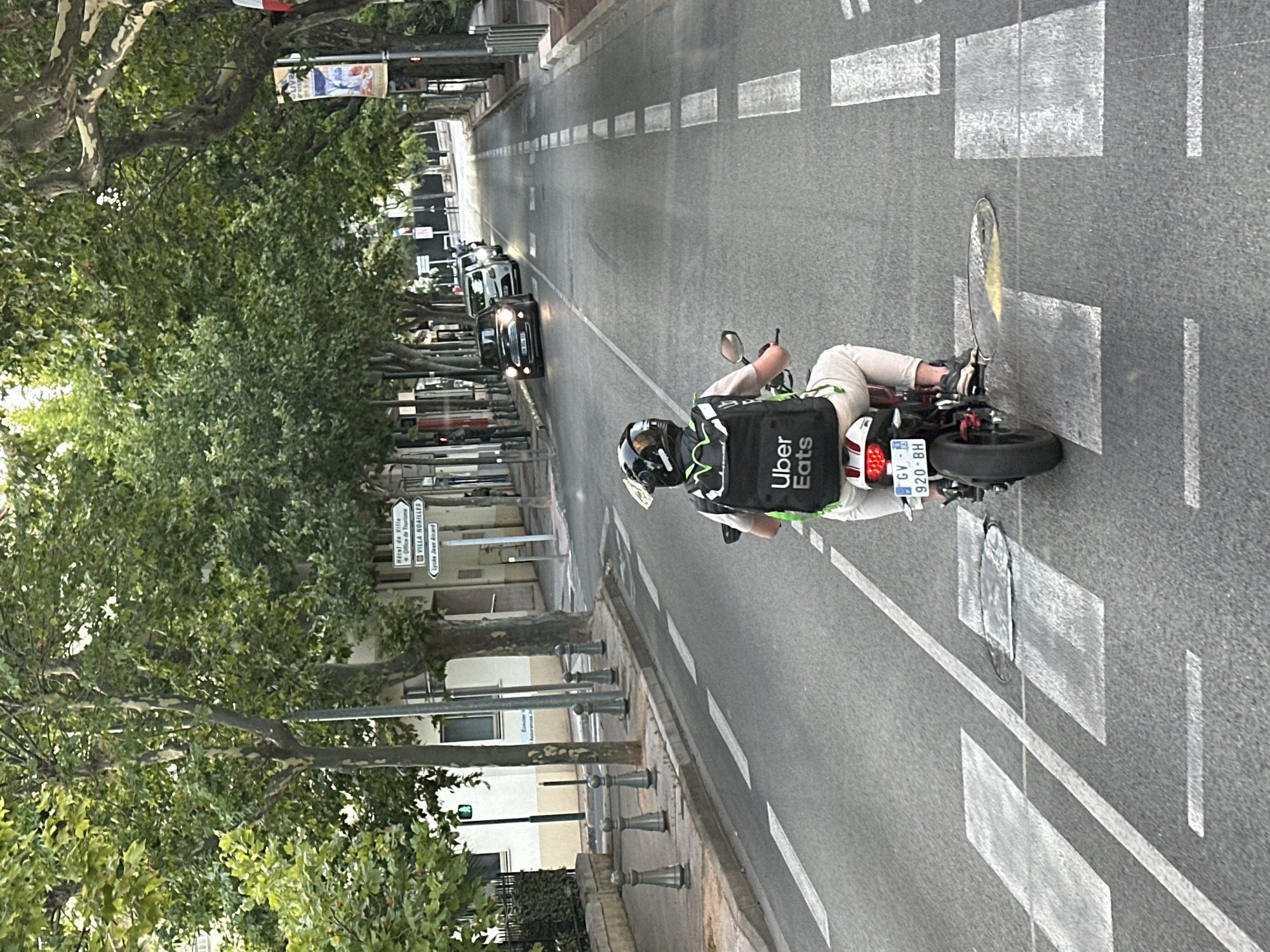 In Hyères, France, an Uber Eats delivery person is seen from behind riding a motorcycle down a tree-lined street. The delivery person is wearing a black helmet with green accents, a tan shirt, and light-colored pants, with a black Uber Eats delivery bag on their back. The motorcycle has a French license plate that reads "GV-320-BH". The street has pedestrian crosswalks and is lined with buildings and trees. Several cars are visible in the distance, driving in the same direction. Traffic lights and street signs, including one indicating "Hotel de Ville - Office de Tourisme" and "Villa Noailles", are present. The scene appears to be in the daytime, with ambient sunlight filtering through the trees.