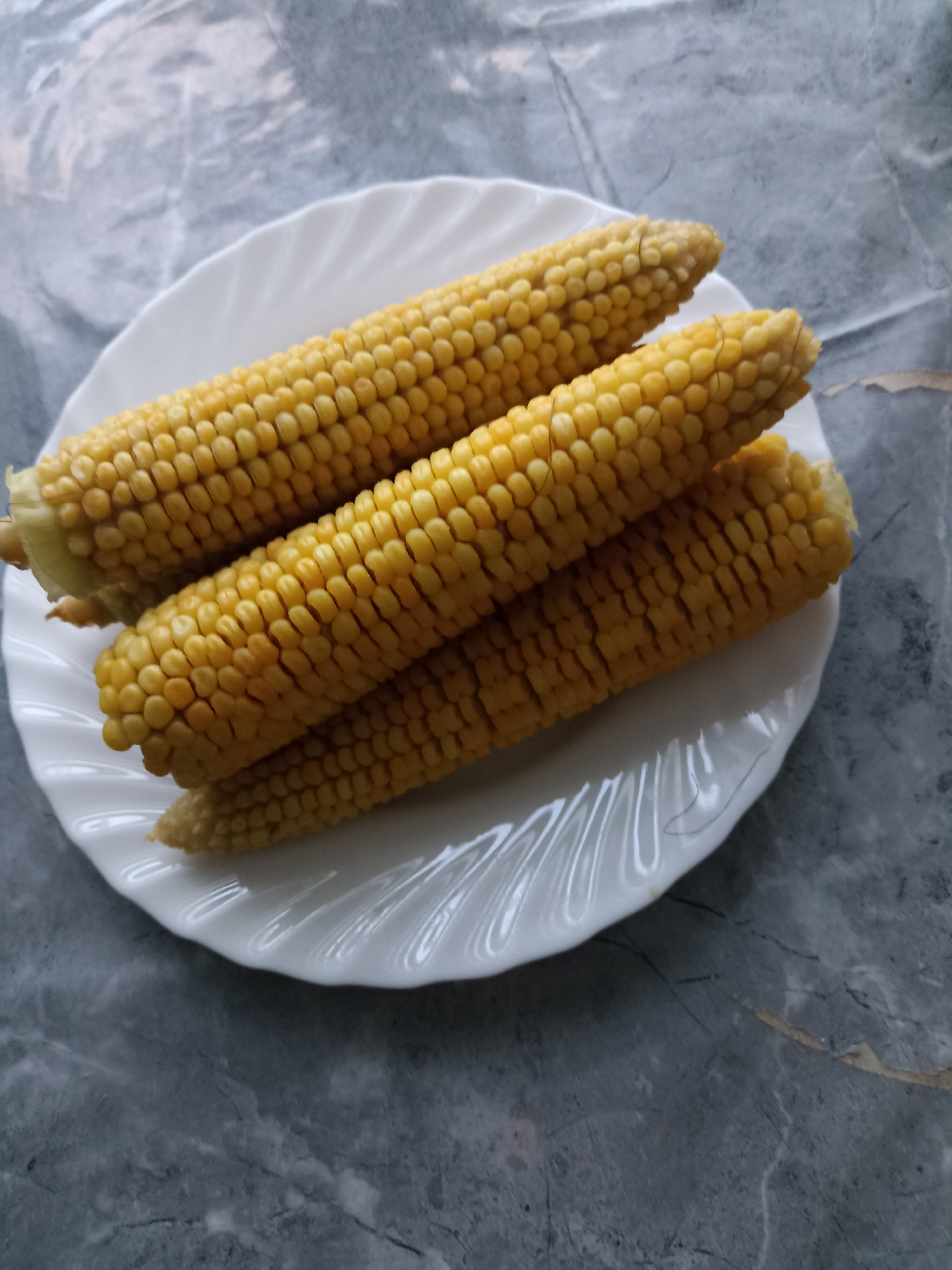 Four ears of corn are arranged on a white, fluted plate. The corn is golden yellow with visible kernels in rows. The plate is positioned on a dark gray, marbled surface. The scene is illuminated from above, casting soft shadows. The image does not contain any people, actions, or interactions. The location in the city of Заринск, Russia, is not visually discernible from the provided image.
