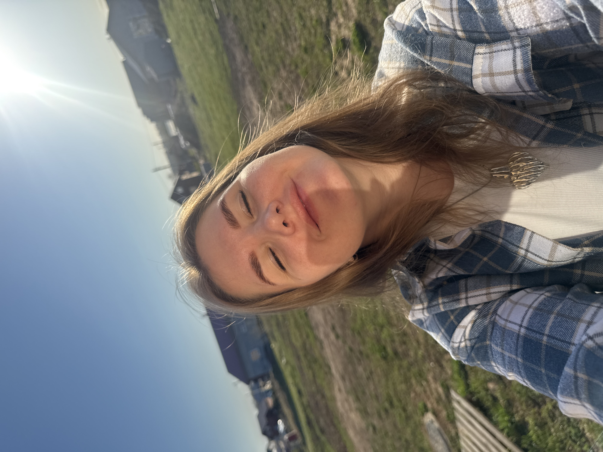 A woman is in Ферма, Russia. She is standing outside with her eyes closed. The sun is shining. She is wearing a white ribbed shirt with a buttoned blue and white plaid shirt over it, and a gold colored pendant necklace with geometric design. The background consists of a blue sky, green grass, a dirt path, some small buildings, and a fence.