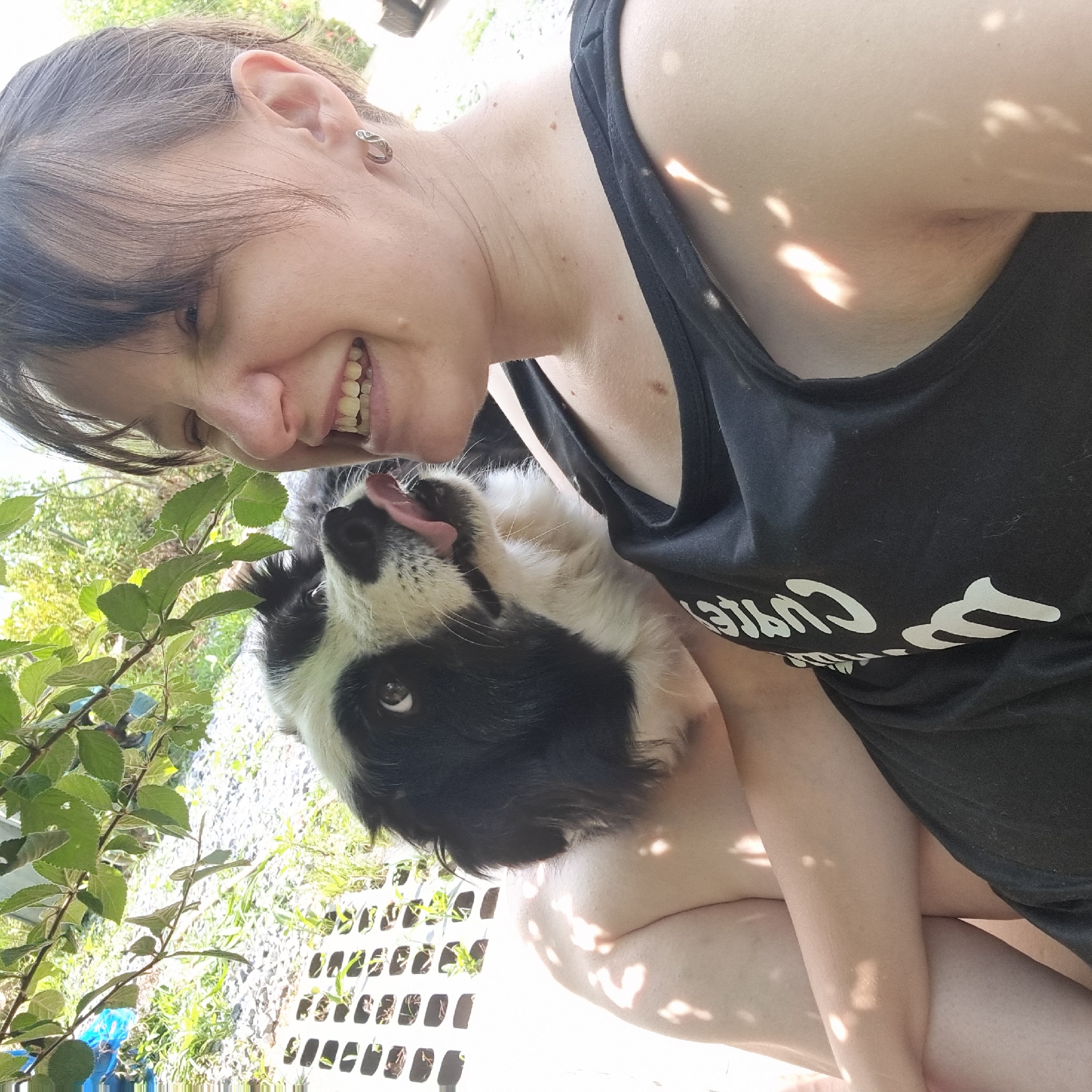 In Dunnae-myeon, South Korea, a young woman with short, dark hair and a wide smile is taking a selfie outdoors. She is wearing a black tank top with white lettering. A black and white Border Collie is nuzzling her face, with its tongue out, appearing to lick her. They are seated outdoors, with green leaves and branches overhead casting dappled shadows on them. In the background, a white slatted structure is visible, along with grass and gravel.