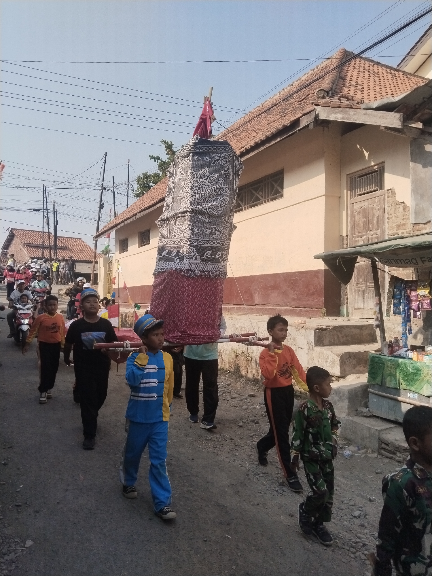 In Jembayat, Indonesia, a group of children are participating in a parade. A large, cylindrical object, possibly a ceremonial item or effigy, is being carried on poles by several boys. The object is draped in patterned fabric, featuring floral motifs in grey and white against a red background. Some of the children are wearing uniforms. One boy in the foreground wears a blue and yellow uniform with a cap. Another boy to his right is dressed in a camouflage military-style uniform. Behind them, other children in casual clothing walk along a paved road lined with houses and power lines. A motorcycle is visible further back in the procession. The buildings have traditional tiled roofs and some appear weathered. A small roadside stall selling various items is situated to the right of the frame. The sky is clear and blue, indicating a sunny day.
