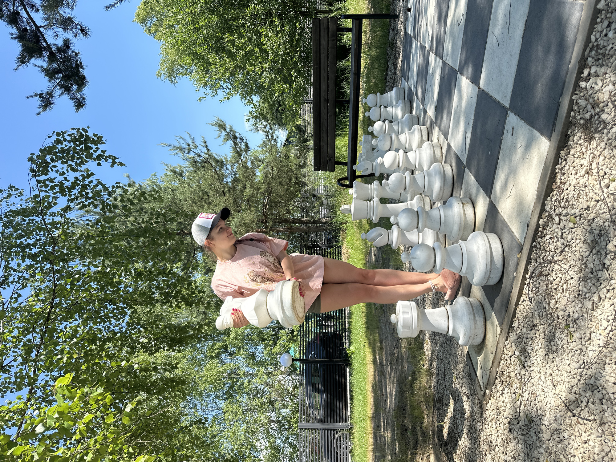 In Mikhalevo, Russia, a young woman with a baseball cap and a pink t-shirt is standing on a large outdoor chessboard. She is holding a white chess piece, likely a queen or king, and is looking down at the board. The chessboard is made of black and white tiles and is surrounded by gravel and grass. In the background, there are trees, a fence, a bench, and a building. The sky is blue and clear, indicating a sunny day.