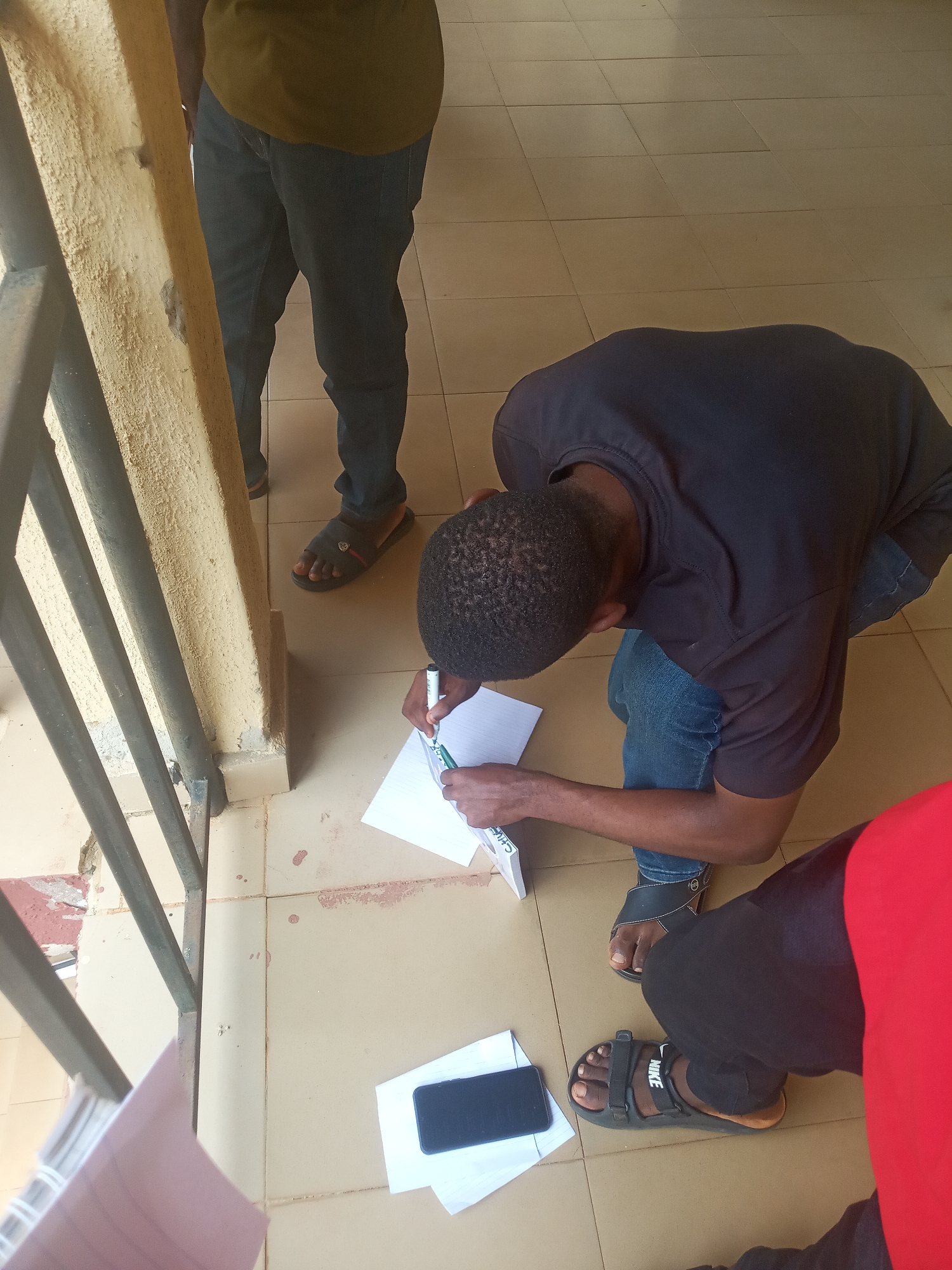 The image captures a ground-level view of several individuals gathered in what appears to be a semi-outdoor or covered area, likely in Nkwelle, Nigeria. The flooring consists of light brown square tiles.
In the foreground, a person wearing a black long-sleeved shirt and blue jeans is crouched down, intently writing on a white sheet of paper using a green and white pen. The word "CHIEF" is visible on the side of the pen. A dark smartphone rests on another piece of paper on the tiled floor near this person. They are wearing black sandals.
To the left, another person stands, with only their lower body visible, clad in dark jeans and dark sandals, positioned next to a weathered light yellow concrete pillar and black metal railings. These structural elements suggest a building's exterior, an open corridor, or a balcony area. In the bottom right corner, the legs of a third person are visible, wearing black pants and black Nike sandals, with the "NIKE" logo clearly visible on the strap. A piece of red fabric or clothing is partially seen near this person.
The scene is illuminated by natural light, indicating it is daytime. The individuals appear focused on the task at hand, suggesting an activity involving document completion or note-taking in a casual setting. Some minor red stains and dirt are visible on the tiled floor near the pillar.