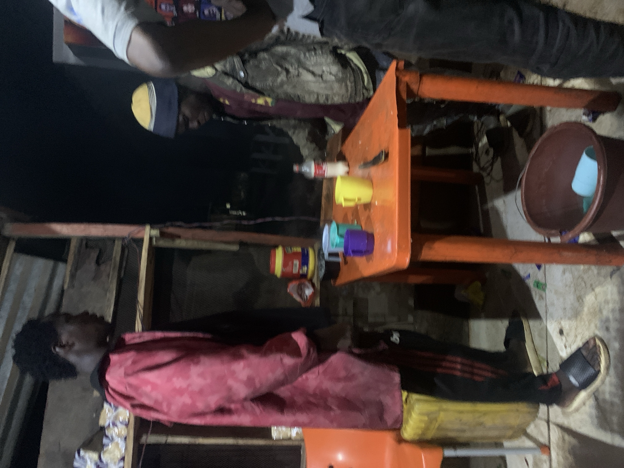 A group of people are gathered around an orange plastic table at night in Gura Top, Nigeria. The lighting appears to be artificial, casting long shadows and creating a dim atmosphere.
On the table, there are several items: a bottle of water with a red label, a yellow mug, a purple mug, and a green mug. A knife is also visible on the table.
One person, wearing a red jacket with a camouflage pattern and black pants with red stripes, stands on the left side of the image. Another person, wearing a yellow and blue beanie and a patterned jacket over a maroon shirt, is seated at the table. A third person, partially visible on the right, is wearing a light-colored shirt and dark pants.
In the background, shelves are stocked with what appear to be bags of food items. A yellow plastic container is situated next to an orange chair on the left. On the floor, a brown bucket contains a blue cup and a green object. The ground appears to be a dirt or concrete surface, possibly wet in places.