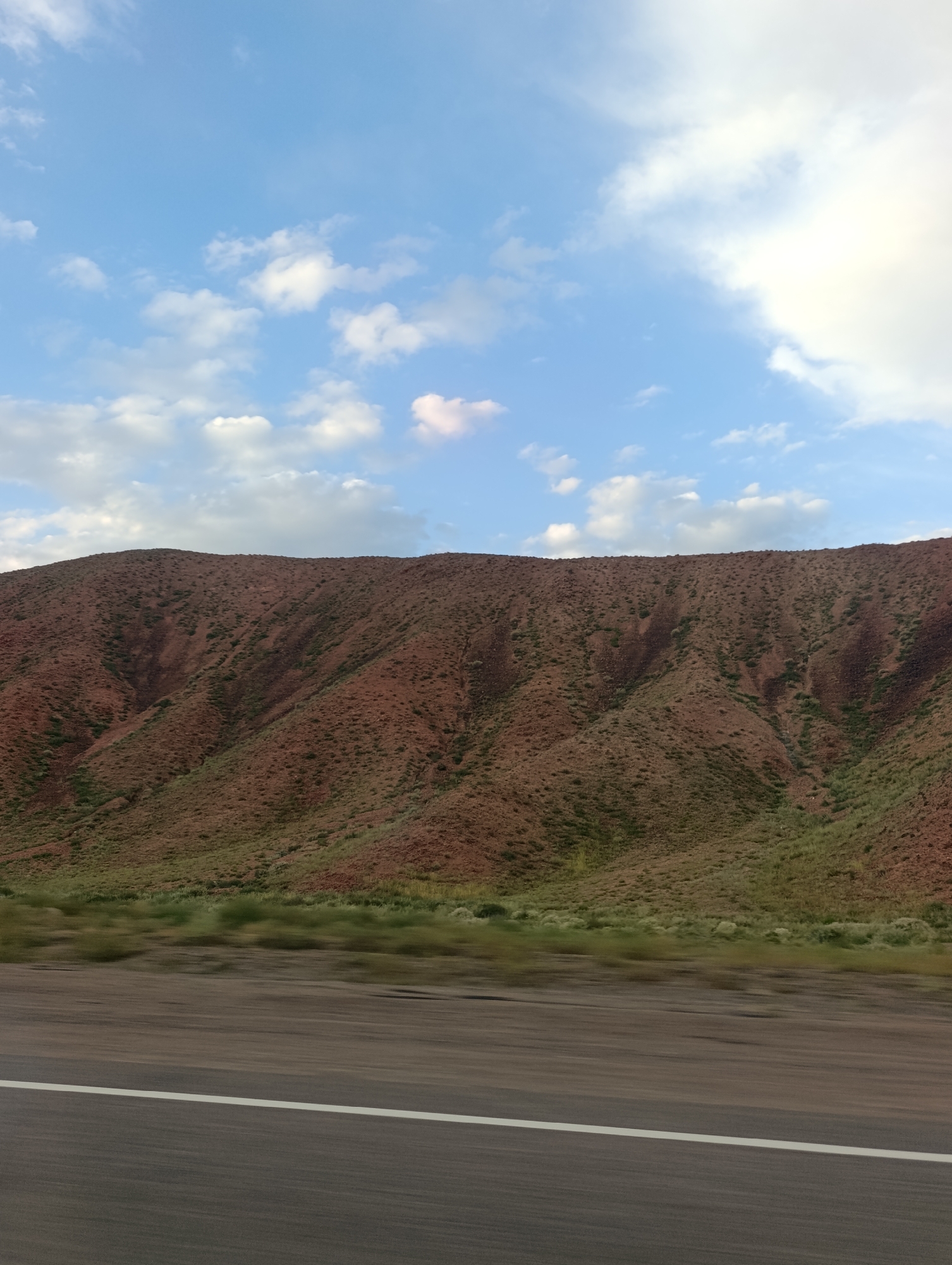 The image captures a natural landscape with a road in the foreground and a mountain in the background, under a sky with scattered clouds. The mountain has a reddish-brown hue with patches of green vegetation, suggesting a somewhat arid environment. The road is paved, with a white line marking a lane, and shows motion blur, indicating the photo was taken from a moving vehicle. The sky is a light blue with white and gray clouds, some of which are wispy and others more solid. No people or specific landmarks are visible, making it difficult to pinpoint an exact location within Orto Tokoy, Kyrgyzstan, beyond it being a general outdoor scene in that region.