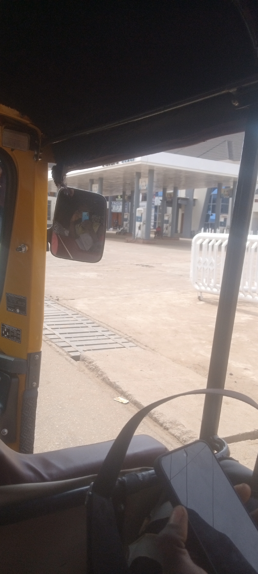 The image depicts a view from inside a yellow vehicle, possibly an auto rickshaw, in Sabon Tasha, Nigeria. The vehicle is at a gas station, evidenced by gas pumps and a station building in the background. A side mirror is visible, reflecting the surrounding environment. There are white barriers in the foreground near the gas station. A person is visible in the bottom right corner holding a phone. The road surface appears to be concrete.