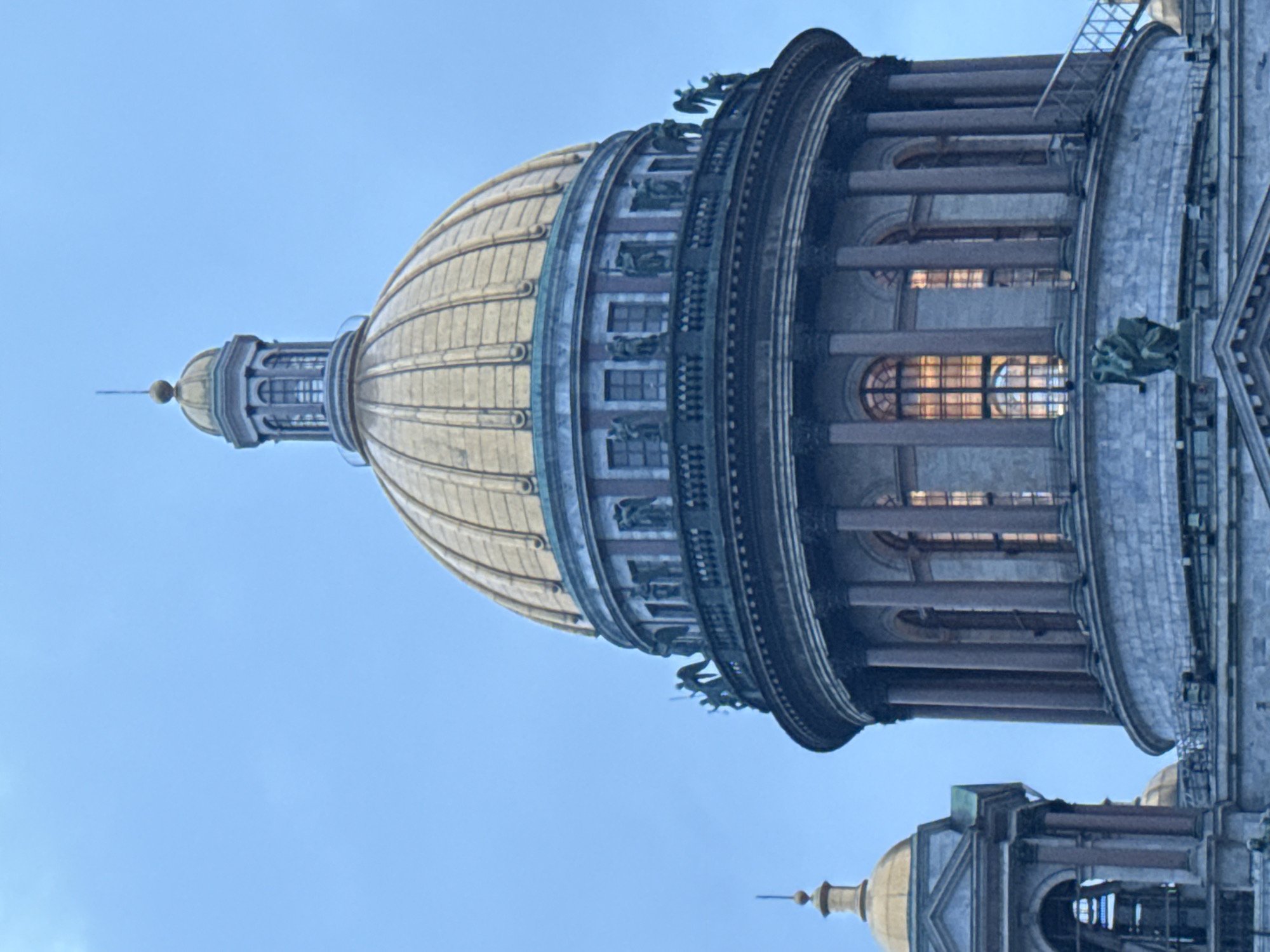 The image shows the golden dome of Saint Isaac's Cathedral, a prominent landmark in Saint Petersburg, Russia. The dome is ornate and detailed, with a lantern structure at its apex. Below the dome, there is a circular drum with windows and decorative sculptures of angels. The building is constructed of stone and features classical architectural elements such as columns and arches. To the left of the main dome, a smaller golden dome and part of a bell tower are visible. The sky is a clear, pale blue, suggesting it might be daytime with overcast conditions. The lighting indicates that the sun is not directly illuminating the scene. The overall impression is one of grandeur and historical significance.