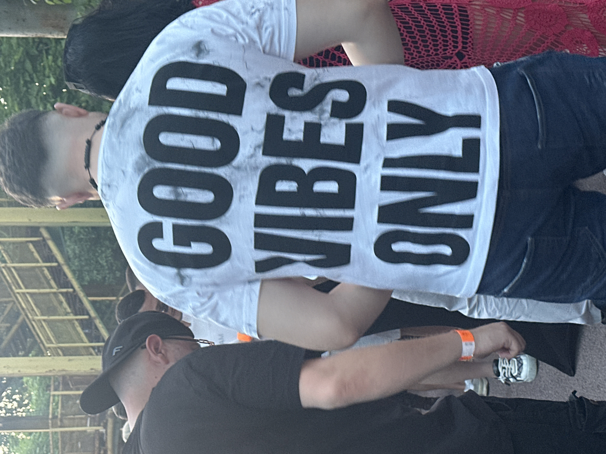 The image shows the back of a man wearing a white t-shirt with the phrase "GOOD VIBES ONLY" in large black letters. He is wearing dark jeans and a beaded necklace. Behind him, a woman is wearing a red crocheted top. In front of him, another man is wearing a black t-shirt and a black baseball cap with a white "F" on it. The man in black has a wristband on his arm and is standing near a pair of white and black sneakers. The scene appears to be outdoors, possibly at a festival or gathering. The background includes a metal structure resembling a partially obscured roof. There is no definitive visual evidence to confirm the location within Plovdiv, Bulgaria.