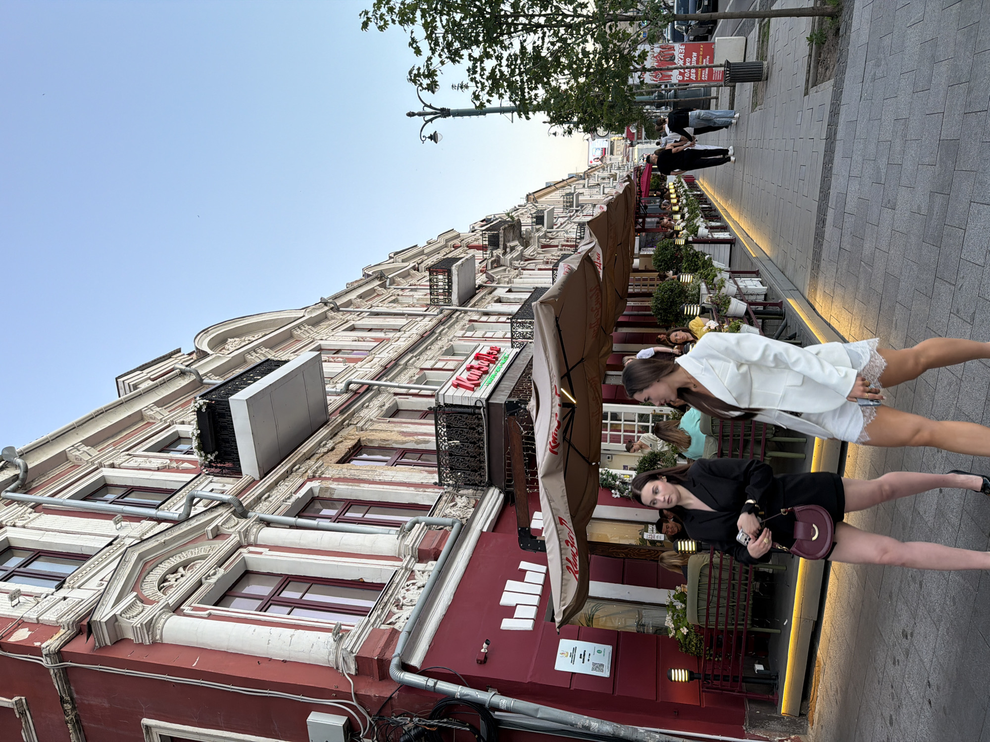 This image depicts an outdoor scene on a street lined with buildings and cafes. The buildings are tall with ornate architecture, featuring red and white facades, balconies, and numerous windows. In the foreground, two young women are walking on the sidewalk. One woman is wearing a white blazer and a short skirt, while the other is in a black dress and carrying a burgundy handbag. They appear to be engaged in conversation or simply walking together.
Several cafe tables with umbrellas are set up along the sidewalk, suggesting a dining or social area. In the background, more people can be seen walking on the sidewalk and sitting at tables. The scene is illuminated by natural light, indicating it is daytime, likely late afternoon or early evening given the warm light and shadows. The sky is clear and blue.
A street lamp with an old-fashioned design is visible among the trees lining the sidewalk. There is some visible text on signs attached to the buildings, one of which appears to be a restaurant name. The overall atmosphere seems relaxed and casual, typical of a European city street with outdoor seating. The location is identified as Novaya Usman', Russia.