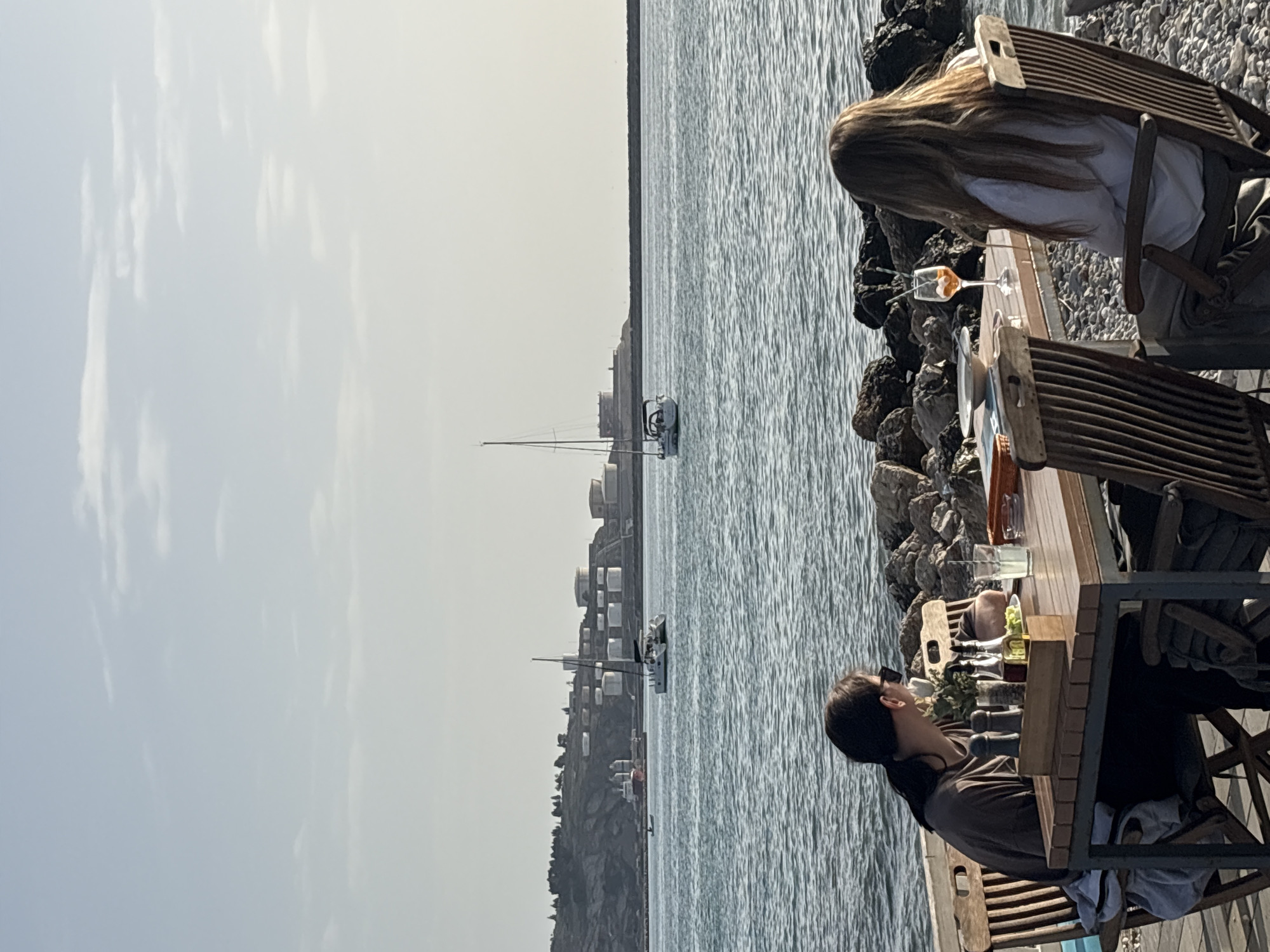 Two people are seated at a wooden table in an outdoor dining area, seemingly at a restaurant, in Bar, Montenegro. The restaurant is located directly next to the coastline of the Adriatic sea. The individuals are positioned next to each other, facing the body of water. The person closest to the camera is dark-haired and wears dark sunglasses. The person farthest from the camera has long blonde hair. The table holds beverages and food items, including a partially consumed Aperol spritz. The sea contains several boats. Beyond the sea, land is visible, including buildings that appear industrial in nature. The sky is hazy. The image is captured from a slightly elevated position and appears to be rotated to the right. The landscape in the distance features buildings and greenery.