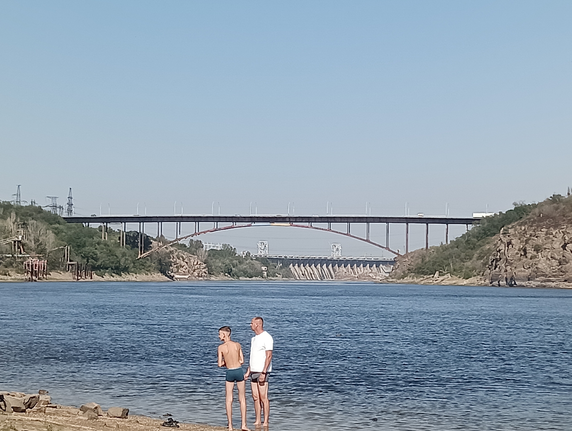 Two individuals, an adult male and a younger male, stand partially submerged in the shallow water at the edge of a wide, blue river. The adult, wearing a white t-shirt and dark swim trunks, faces left. The younger male, shirtless and wearing dark swim trunks, faces forward and slightly left, looking at the adult. They are positioned on a gravelly shoreline. In the background, a large, arched bridge spans the river, supported by numerous piers. Further in the distance, another bridge or dam structure is visible, with a series of tiered walls. The surrounding landscape is hilly and covered with vegetation on the far banks of the river. The sky is clear and light blue. The scene appears to be outdoors in a natural setting, likely a riverfront area.