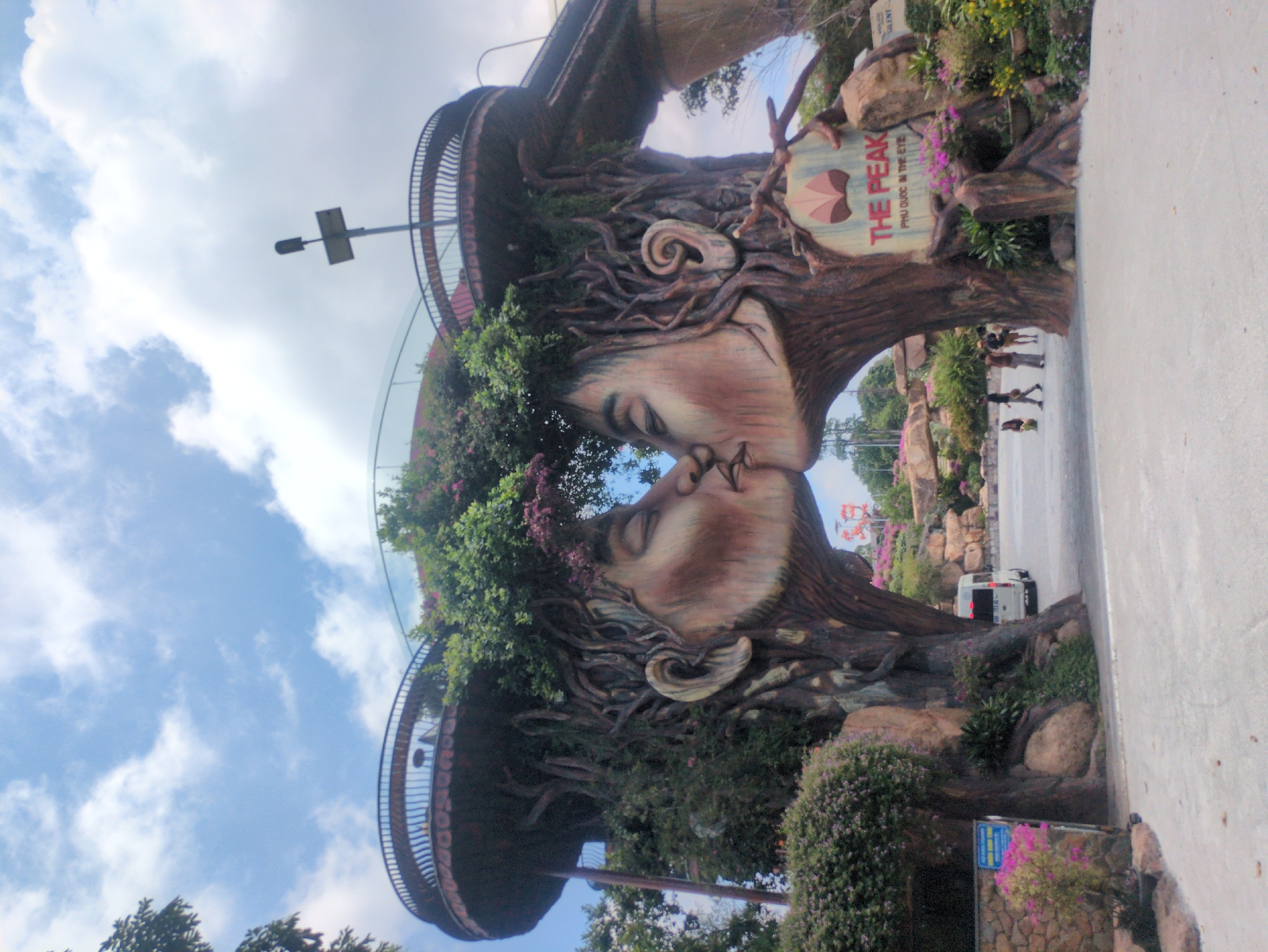 The image shows a large art installation titled "The Peak, Phú Quốc In The Eye" featuring two faces constructed of wood and plants facing each other as if kissing. It is located in Phú Quốc, Vietnam. The faces are placed on top of what appear to be observation platforms with metal railings. The sky is partially cloudy. In the background, there is a road with a white van on it and several people walking in the distance. Plants and flowers grow along the base of the installation.