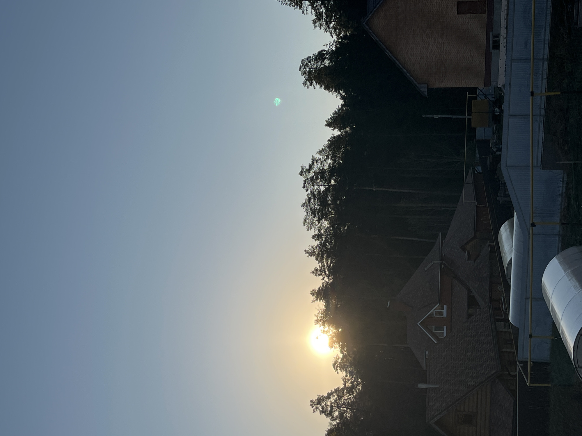 The image depicts a daytime scene in or near Chaikovsky, Russia, as the metadata indicates. The sky, visible in the upper portion of the frame, is a pale blue. The sun is positioned low on the horizon, partially obscured by a dense line of trees. Several buildings, with varied rooflines and construction materials, are visible among and behind the trees. To the right of the frame, cylindrical structures and metal frameworks are prominent. There are no people visible in the image. A green blob is visible in the sky.