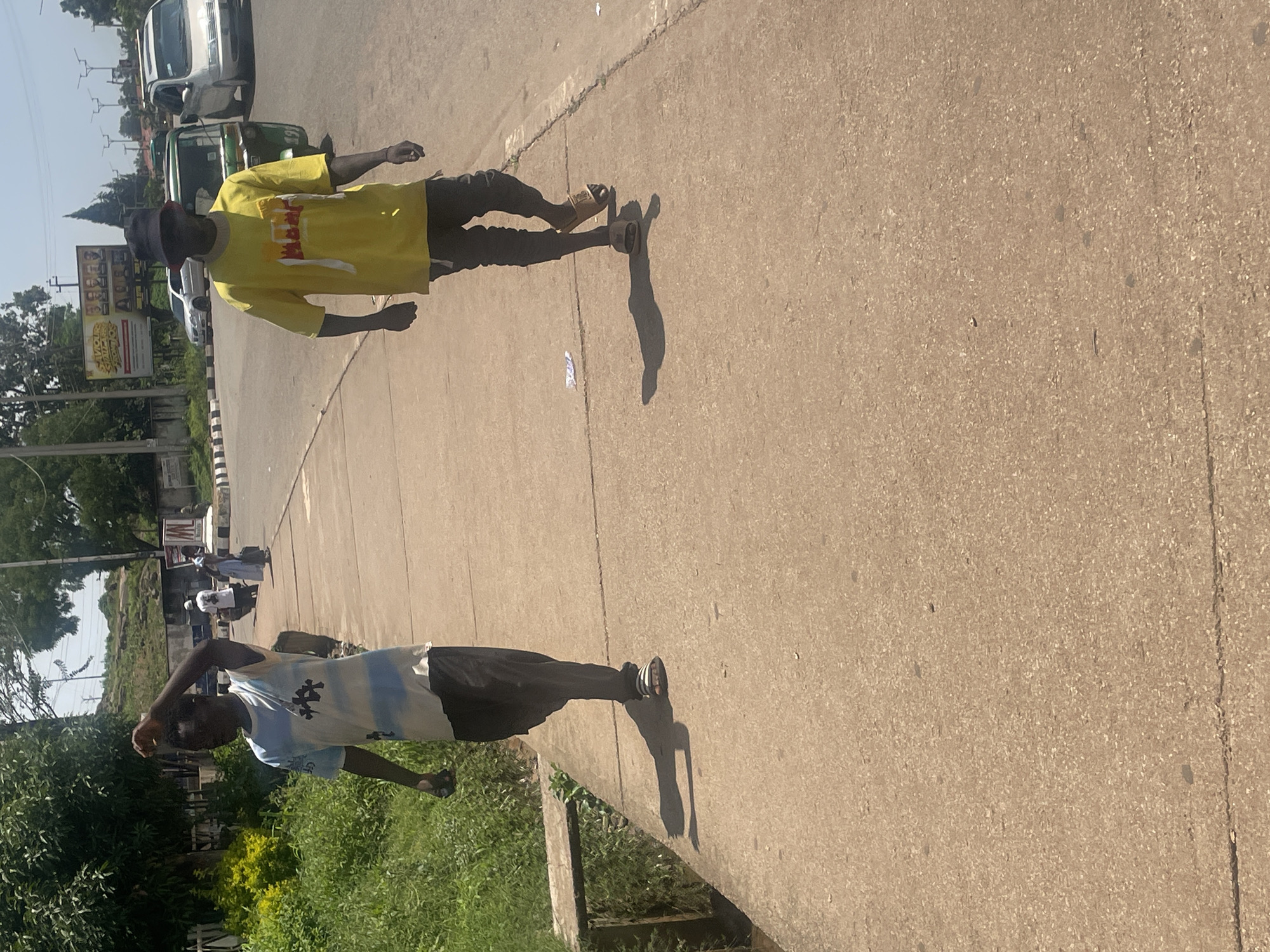 The image depicts a street scene in Gura Top, Nigeria, on a sunny day. Two young men are walking on a concrete sidewalk. The man in the foreground on the left wears a blue and white striped t-shirt and dark pants, with his right arm raised. The man on the right wears a bright yellow t-shirt, a dark bucket hat, and dark pants. Behind them, a green three-wheeled auto-rickshaw, a silver car, and a white car are visible on the road. The environment includes lush green vegetation on the left and a roadside with some distant buildings and utility poles. The sidewalk is well-worn concrete, with some debris visible on its surface. The lighting suggests it is midday, with strong shadows cast by the individuals.