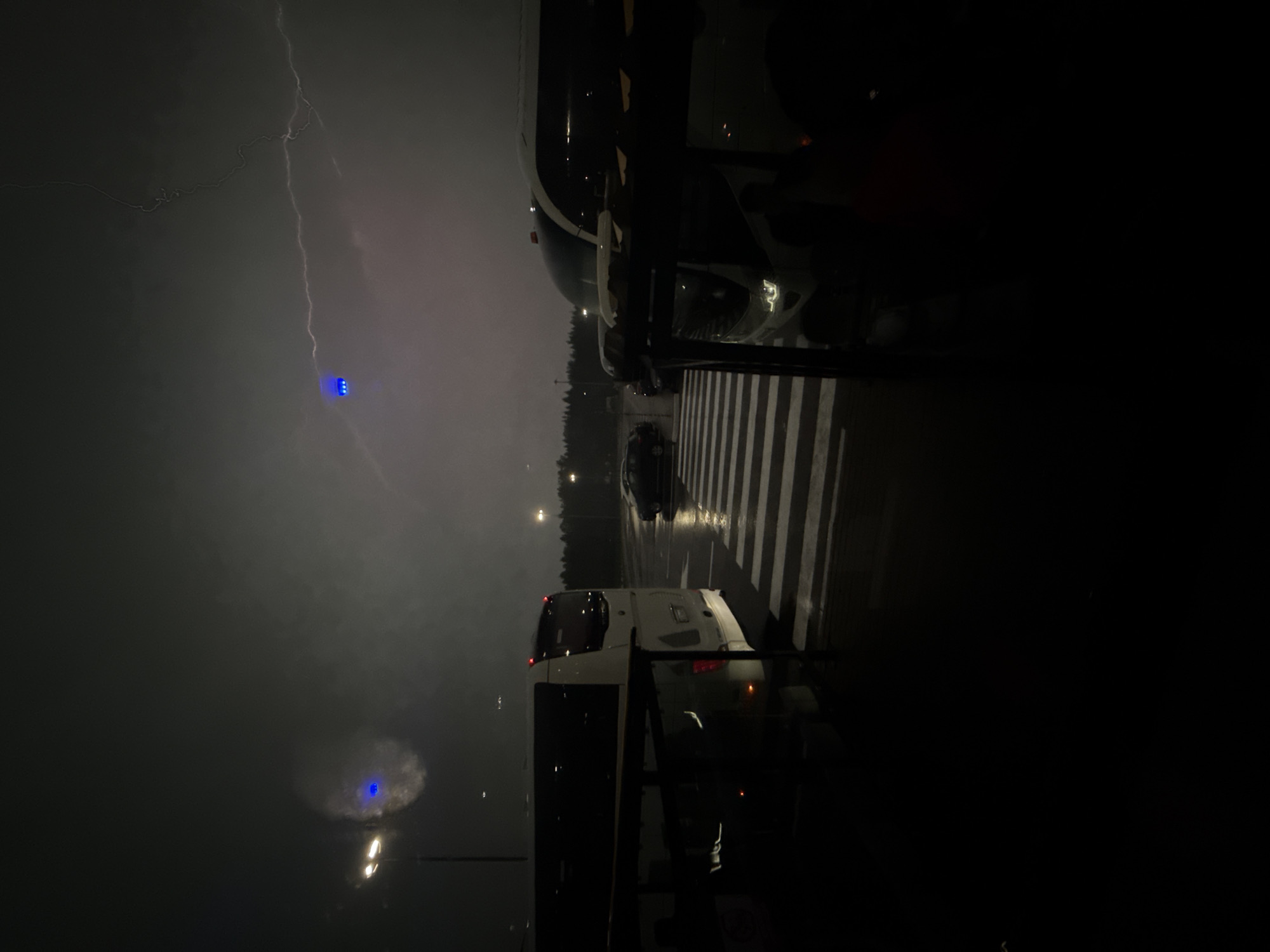The image captures a dark, stormy night at what appears to be a bus parking lot or station in Lupanovo, Russia. A lightning strike illuminates the dark sky, with faint branches of lightning visible. In the foreground, a white bus is parked on the left, partially obscured by a dark structure. Across a wet, crosswalked area, another white bus is parked on the right, with its headlights on. A dark car is visible between the buses, driving across the crosswalk. The ground is wet, suggesting recent rain. Streetlights with blue glows are visible in the distance. The overall scene is dimly lit, with the exception of the lightning and vehicle lights.