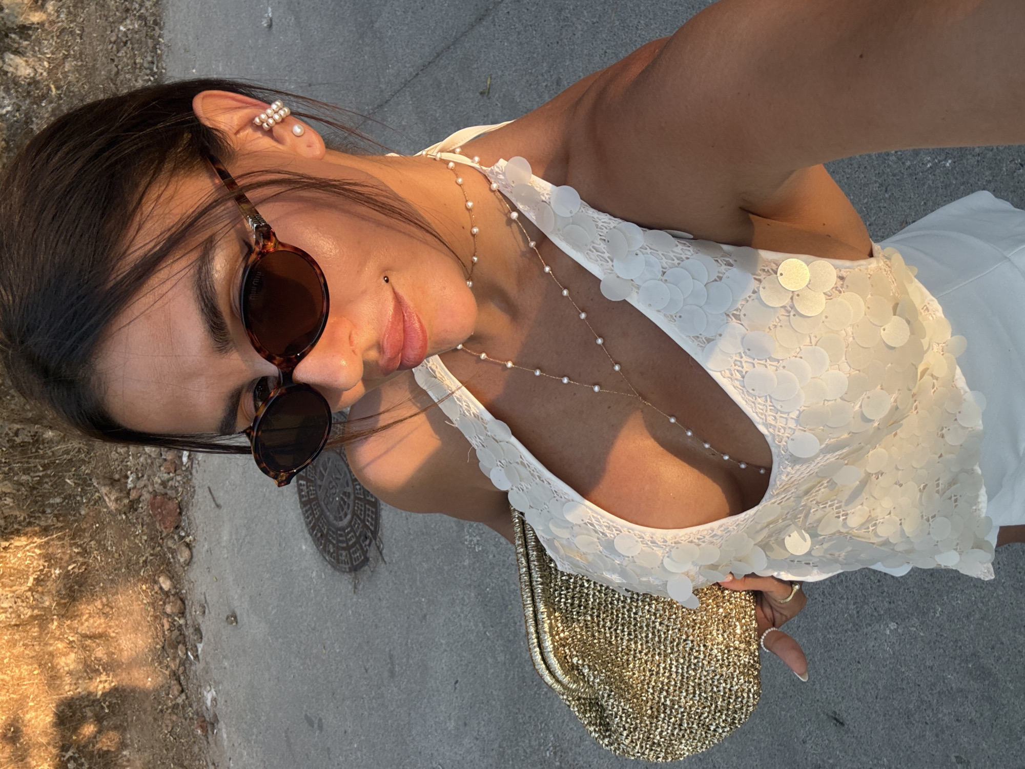The image features a close-up, tilted shot of a woman outdoors. She is wearing sunglasses with tortoiseshell frames and a white, sequined top. A delicate pearl necklace is visible around her neck. The woman has a facial piercing below her lower lip and is wearing pearl earrings on her left ear. She is also holding a gold, textured clutch bag. The setting appears to be a street or sidewalk, with a manhole cover visible in the background and some dry vegetation on the left. The lighting suggests it is late afternoon or early evening, with warm sunlight casting shadows. There are no visible signs with text. The woman's expression is neutral, and the overall mood is casual. The location context provided is Fethiye, Turkey.