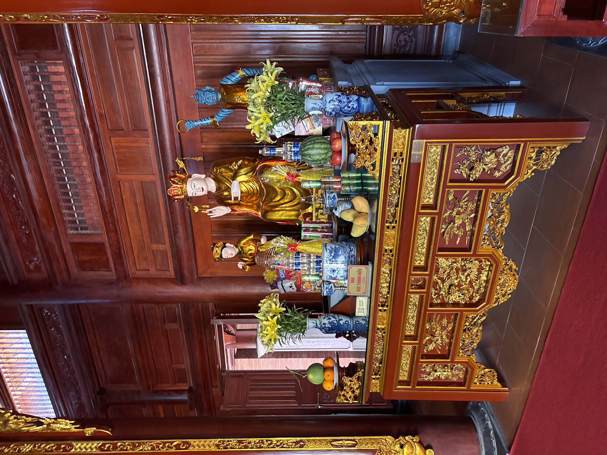 The image shows an altar inside a building in Nam Nha Trang Ward, Vietnam. The altar is made of red wood with elaborate gold detailing. On the altar are statues of figures, including one painted blue, along with offerings of fruit, flowers, and canned goods. The walls and ceiling of the building are also made of reddish-brown wood.