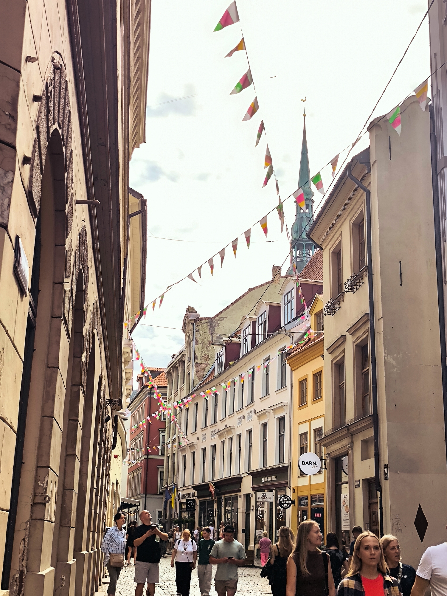 The image shows a narrow street in Riga, Latvia, lined with old buildings and decorated with colorful bunting. A group of people are walking down the cobblestone street, some looking at their phones while others chat amongst themselves. In the background, a green spire rises above the buildings, likely belonging to a church. The architecture suggests a historic European city, consistent with Riga's Old Town. The lighting indicates it is daytime, with some shadows cast by the buildings.