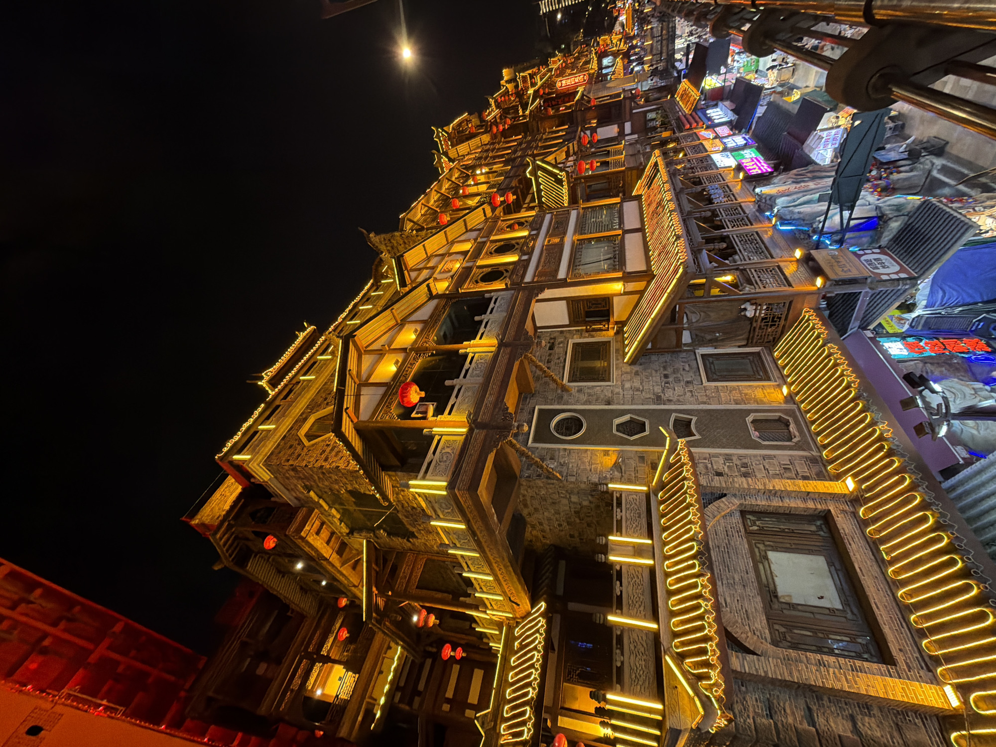 The image depicts a nighttime view of Hongyadong, a multi-story, traditional stilted building complex located in the Yuzhong District of Chongqing, China. The architecture features traditional Chinese design with tiered roofs, intricate wooden balconies, and stone facades. The buildings are illuminated with numerous warm yellow lights, outlining the eaves and balconies, and punctuated by red lanterns. The scene captures the lively atmosphere of the district, with various shops and stalls visible on lower levels, indicated by colorful signage. A walkway or bridge is present in the foreground, offering a vantage point of the bustling street below. The sky is dark, highlighting the illuminated structures. There are no discernible individuals in the immediate foreground or midground in focus.