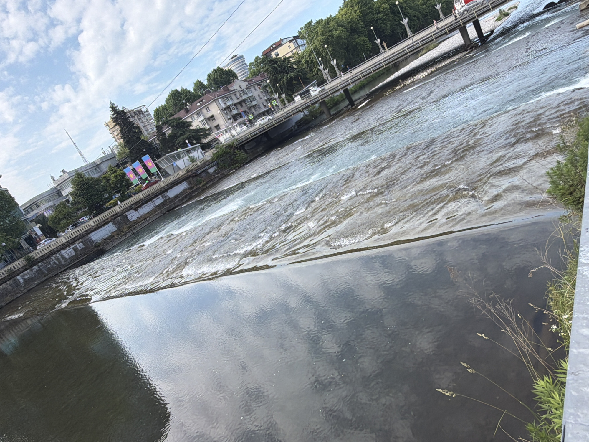 The image captures a slightly tilted, wide-angle view of a river flowing through a city, likely Sochi, Russia. The foreground is dominated by the dark, reflective surface of the river, with ripples and a faint reflection of the sky and clouds. In the middle ground, the river transforms into a series of rapids, with whitewater churning over submerged rocks.
Along the riverbank, there's a concrete embankment. Beyond the embankment, a street with cars and buildings is visible. The buildings are varied in style, some appearing to be older, multi-story structures, while others are more modern. A bridge spans the river in the background, with a few pedestrians and vehicles visible crossing it. Trees line the opposite bank of the river, providing a green contrast to the urban landscape. The sky above is blue with scattered white clouds. The overall impression is of a lively urban environment integrated with a natural river system.
