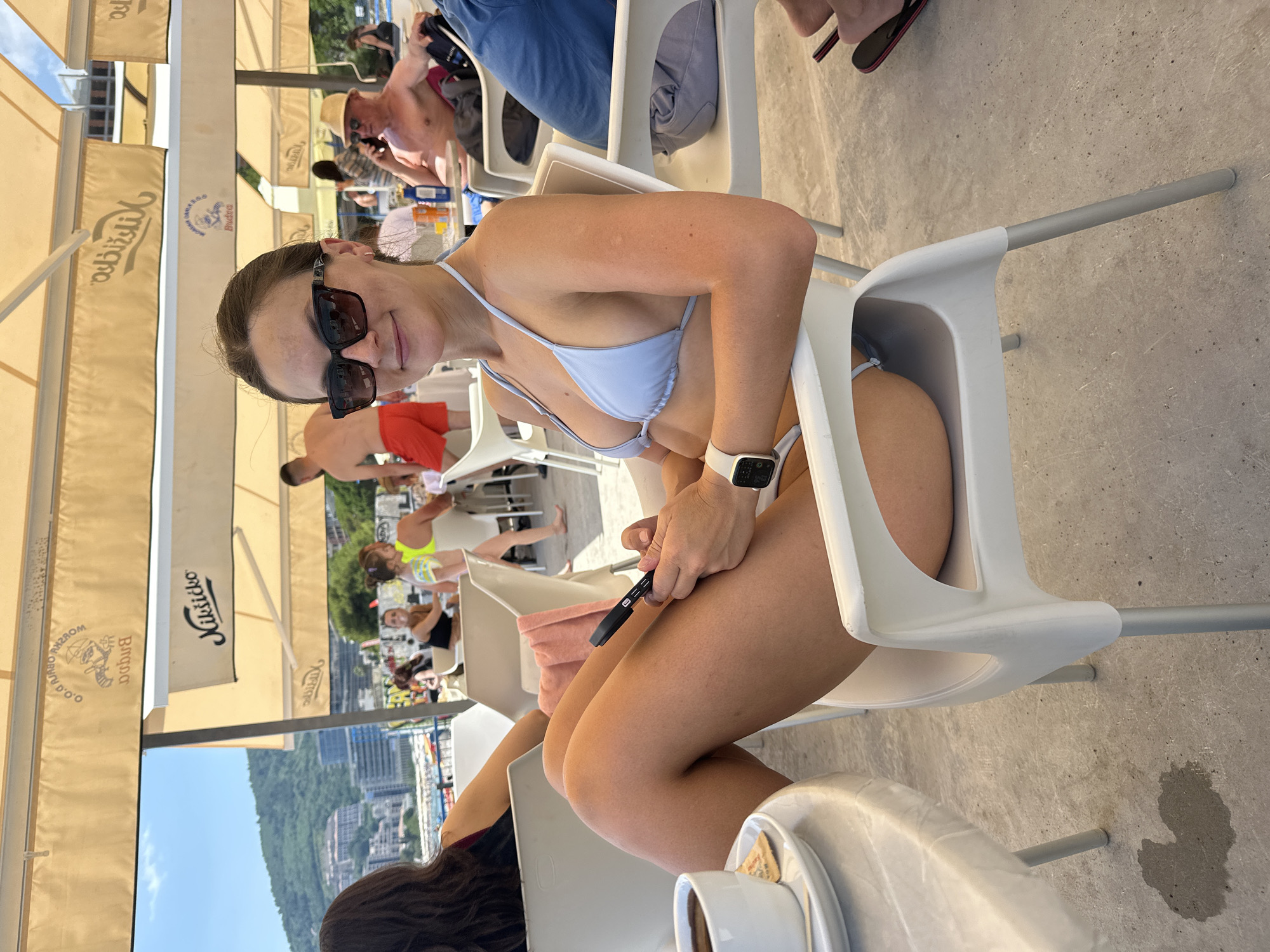 The image shows a woman sitting at an outdoor cafe in Budva, Montenegro. She is wearing a light blue bikini and sunglasses, and she is holding a smartphone. In the background, other people are visible, as well as the cafe's umbrellas and awnings, which are branded with the "Kiliksio" logo. The cafe appears to be located near the coast, with a view of the city and hills in the distance. The ground is concrete, and there is a small puddle of liquid on the ground to the left of the woman's chair. The woman is looking at the camera and smiling.