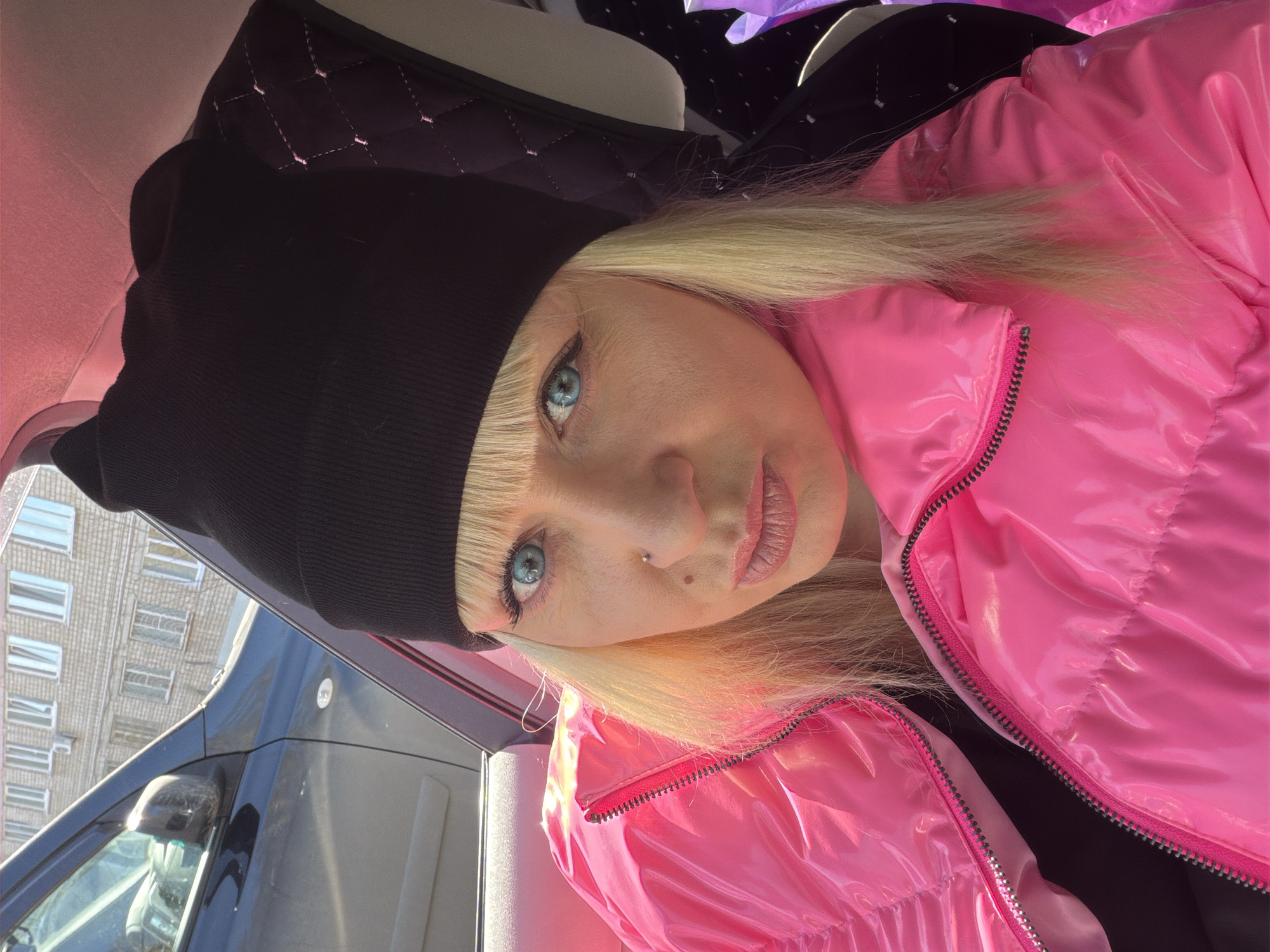 A woman with fair skin, blonde hair, and blue eyes is seen wearing a black beanie and a bright pink, shiny puffer jacket. She has a piercing in her nose and is wearing light pink lip gloss. The background reveals a vehicle window and the corner of a building that is typical architecture in Chaikovsky, Russia.