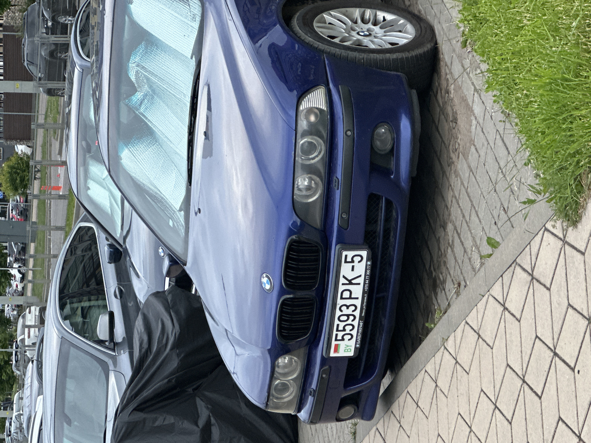 A blue BMW sedan is parked on a paved surface next to a grassy area and a sidewalk. The car has a license plate that reads "5593 PK-5 BY". The front of the car is visible, including the headlights, grille, and bumper. The car is equipped with sunshades in the windshield. In the background, other cars and buildings are visible.
