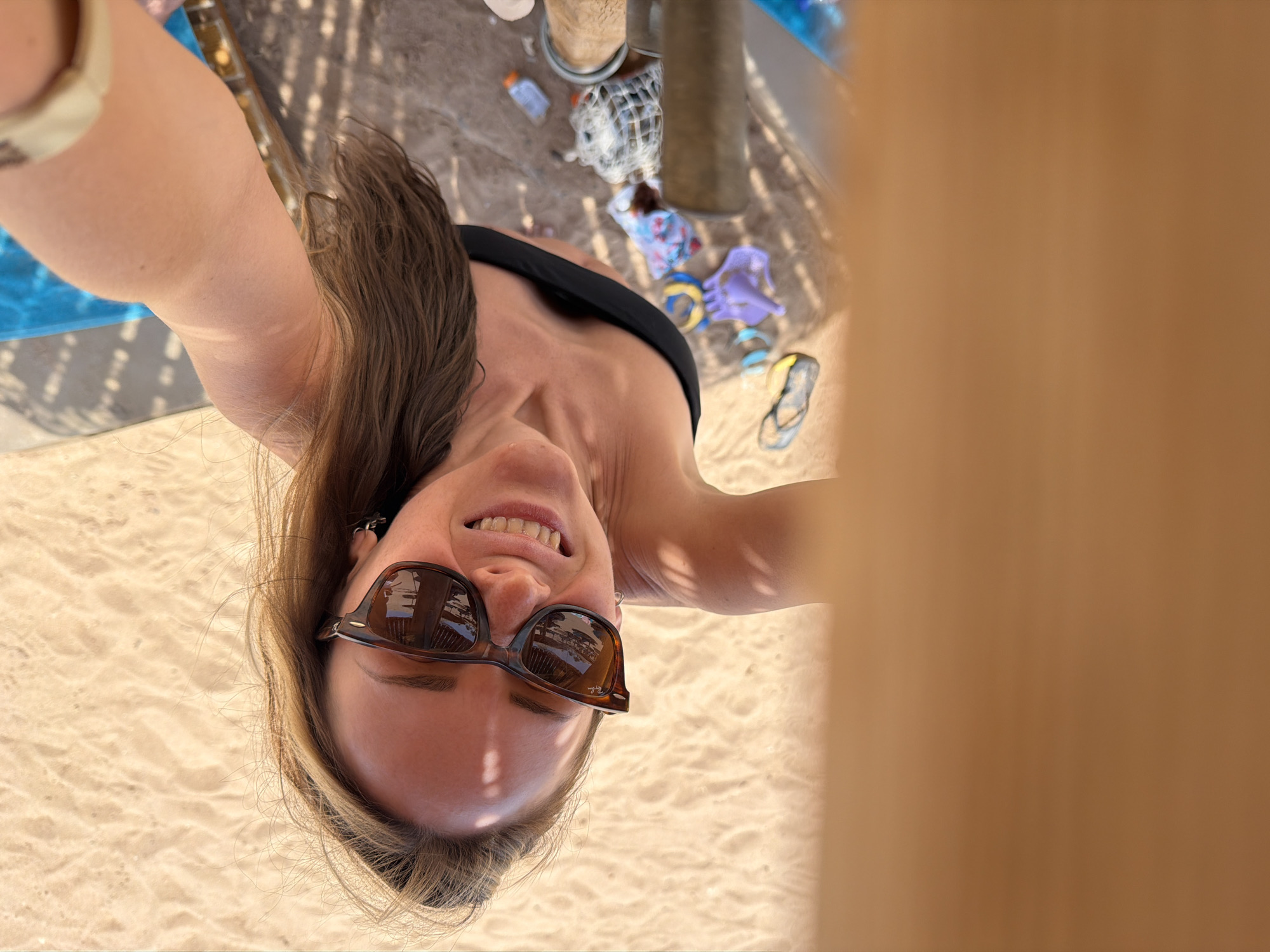 A brightly lit, low-angle selfie captures a smiling woman on a sunny beach, likely in Sharm El Sheikh, Egypt. The image is rotated 90 degrees counter-clockwise, making the woman's head appear towards the bottom left. She has long, light brown hair flowing over her shoulders and is looking directly up at the camera with a wide, happy smile that reveals her teeth. She wears dark, rectangular sunglasses with "Ray-Ban" visible on the right arm, and a black bandeau-style bikini top. Her left arm is visible in the upper left corner.
The background shows a sandy beach floor with clear, dappled shadows, suggesting strong sunlight filtered through a slatted cover or umbrella. Scattered on the sand are various beach items: a blue mat or towel, two light-colored support poles (possibly for a beach umbrella or cabana), a white mesh bag, a small white bottle, colorful children's clothing or toys (including purple fabric and a floral pattern), a yellow ball, and a pair of flip-flops. A large, blurred, light brown structure partially frames the right side of the image, reinforcing the impression of a shaded beach setup. The scene conveys a relaxed and joyful atmosphere on a sunny day at the beach.