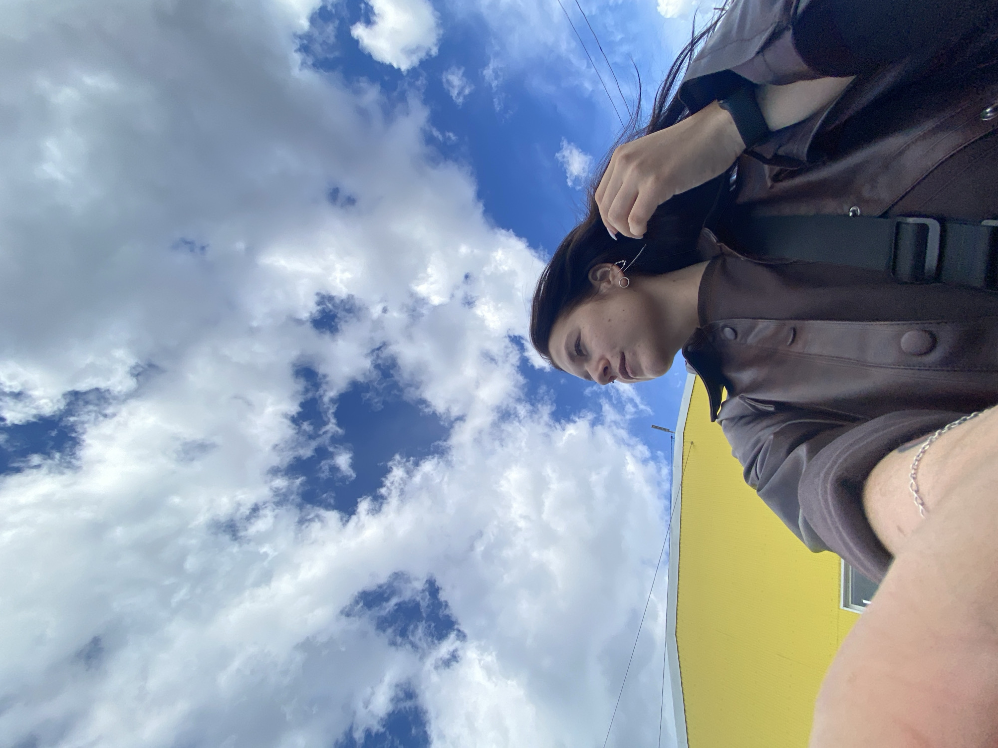 This upward-facing, low-angle shot features a young person positioned on the right side of the frame, looking down and slightly to the left. She has long, dark hair, some strands of which are slightly wind-blown, and wears a dark brown or reddish-brown jacket with visible buttons. A black strap, likely from a bag, crosses her chest. A circular earring is visible on her left ear, and a linear/triangular earring on her right. Her right forearm, adorned with a silver chain bracelet, is partially visible in the bottom right corner.
The dominant background element is a bright blue sky filled with scattered white, fluffy cumulus clouds, suggesting a clear and pleasant daytime. Thin, dark lines, possibly power lines or wires, crisscross the sky in the upper-middle section. A vibrant yellow building or structure occupies the lower right portion of the frame, adding a striking color contrast. The perspective implies a casual outdoor setting, possibly in an urban or suburban environment like Chaikovsky, Russia, where the image is presumed to be taken. No specific activity beyond posing for the photograph is evident, and her expression is neutral. There is no visible text in the image.