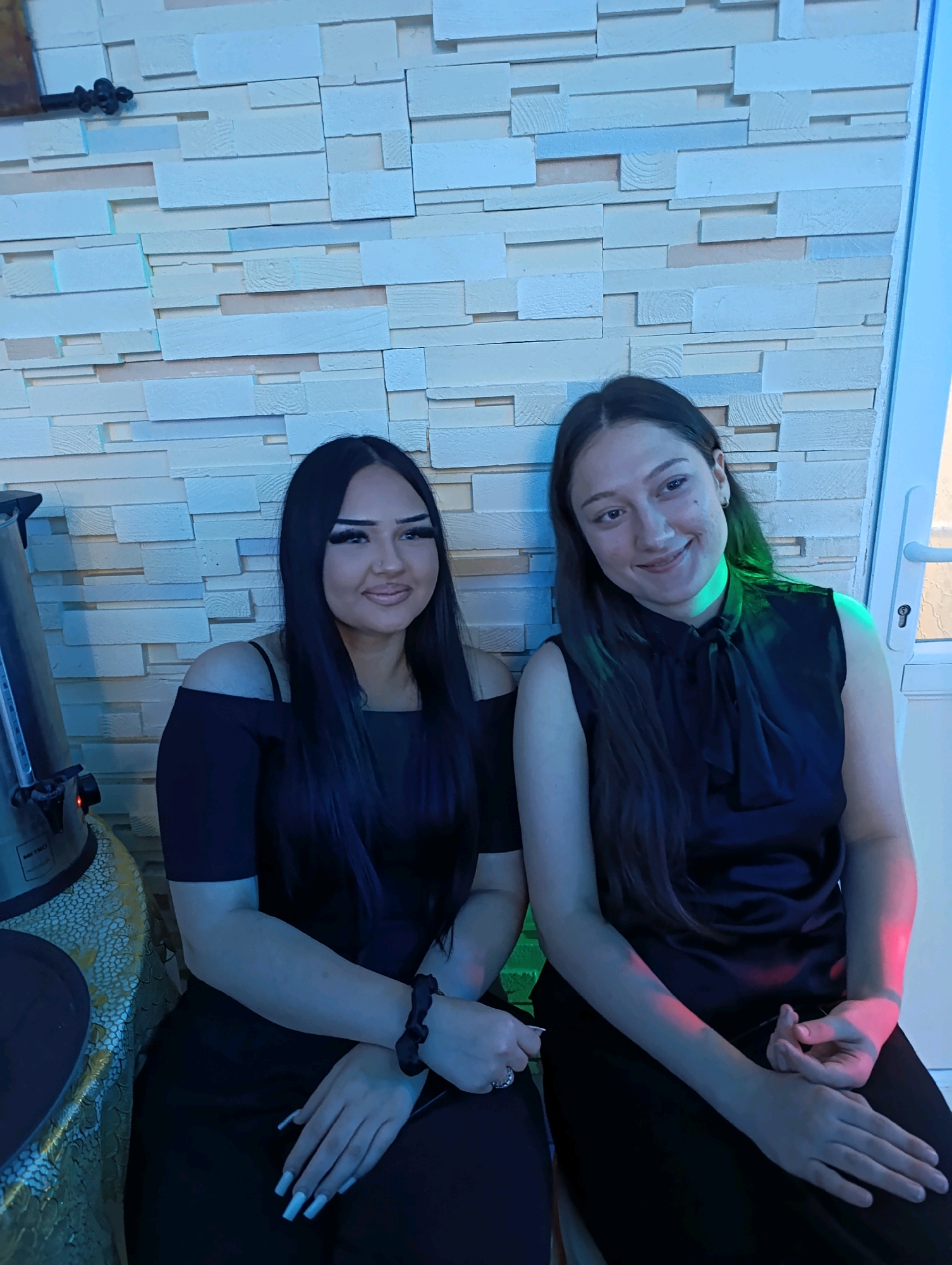 Two young women are seated in front of a textured wall with staggered horizontal wooden panels in varying shades of white and beige. The woman on the left has long, dark hair, pronounced makeup, and wears a black off-the-shoulder top. She is sitting next to a silver-colored appliance with a circular top and a patterned tablecloth beneath it. The woman on the right has dark hair and wears a black sleeveless top with a bow at the neck. Both women are looking towards the camera. The lighting in the scene is a mix of blue and green, with some pink hues present, suggesting a dimly lit environment, possibly a bar or restaurant. The wall treatment and the lighting are characteristic of contemporary interior design. The location is identified as Korzhevo, Moldova.