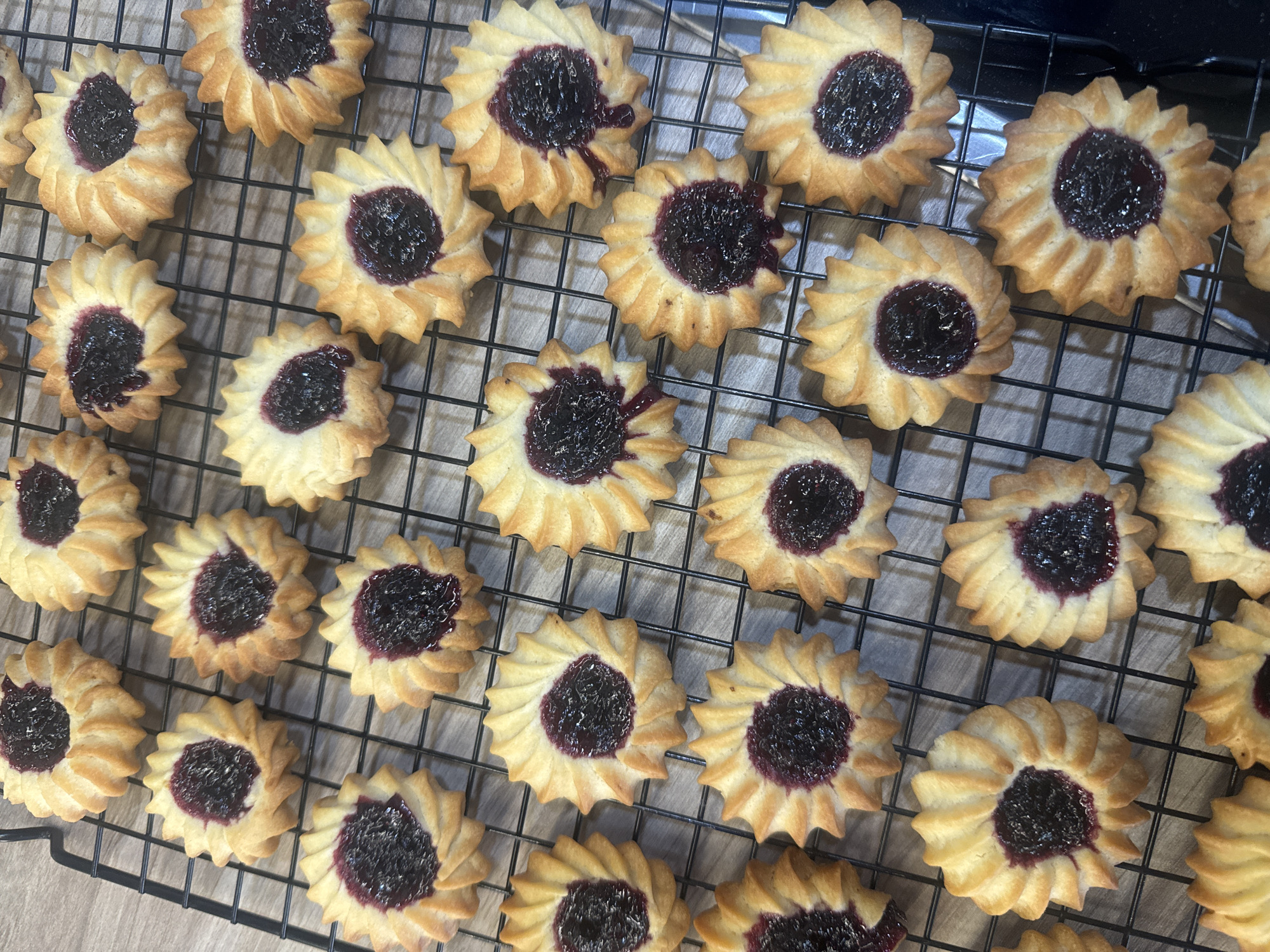 The image shows a cooling rack filled with numerous freshly baked cookies. The cookies are round with a fluted edge, resembling a flower shape. Each cookie is filled with a dark purple jam or filling in the center. They are golden brown and evenly spaced across the black wire rack. The image suggests a home baking environment. The location of Krasnoyarsk, Russia is not identifiable from the visual information provided.